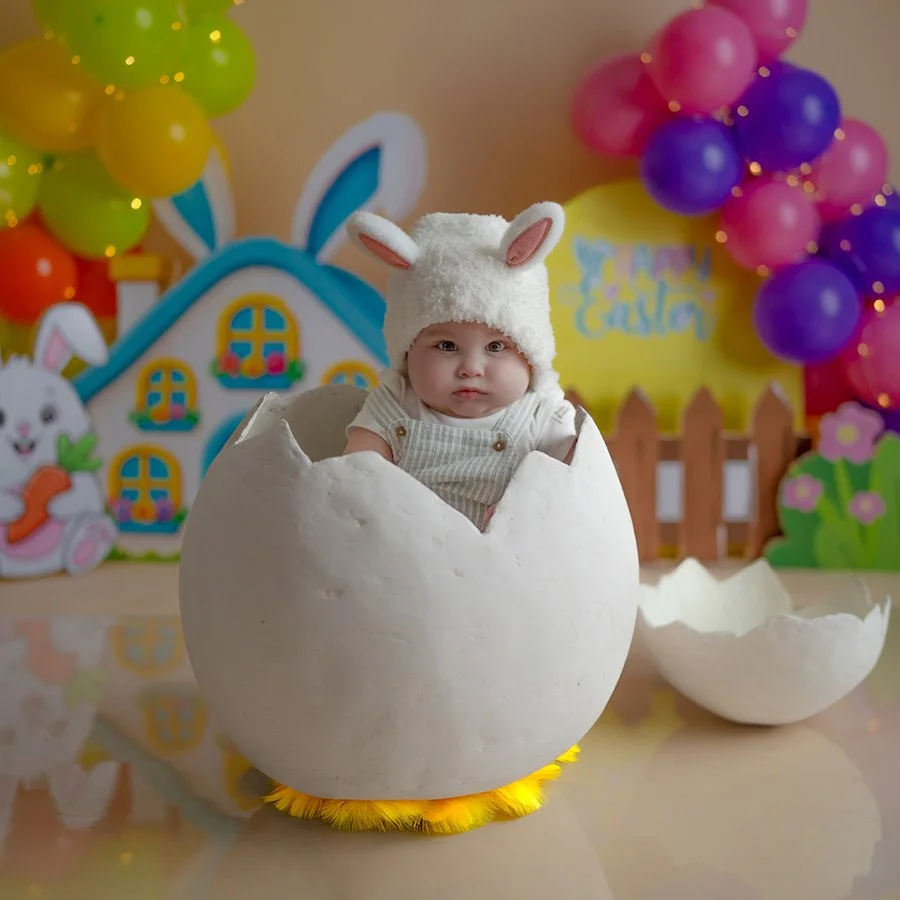 Ein Baby mit Hasenohren-Kappe sitzt in einer Eierschale. Die Dekoration im Hintergrund ist bunt mit Osterelementen.