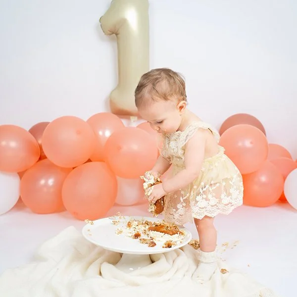 Ein kleines Kind beim ersten Geburtstag, umgeben von rosa und weißen Luftballons, mit einer großen Goldnummer eins Dekoration im Hintergrund.