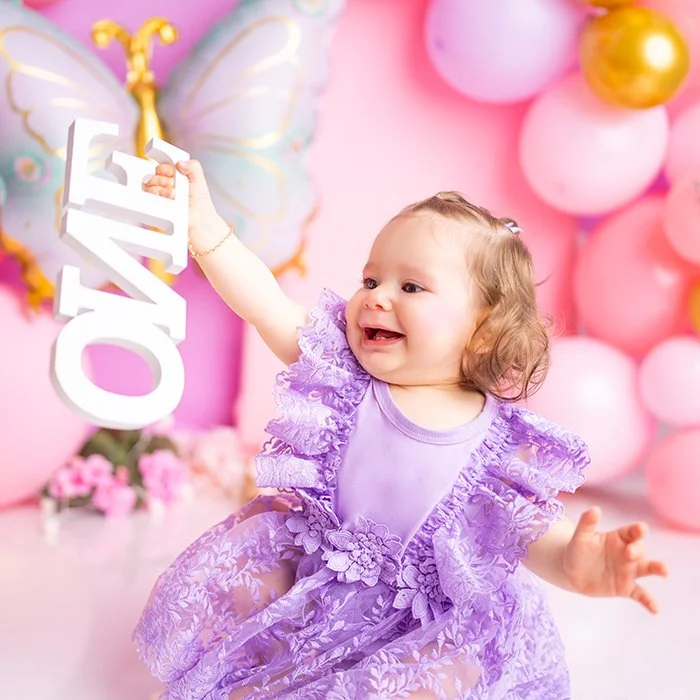 Ein lachendes Baby in einem lila Kleider mit Rüschen, umgeben von rosa und goldenen Ballons, hält ein weißes Schild mit der Aufschrift "ONE".