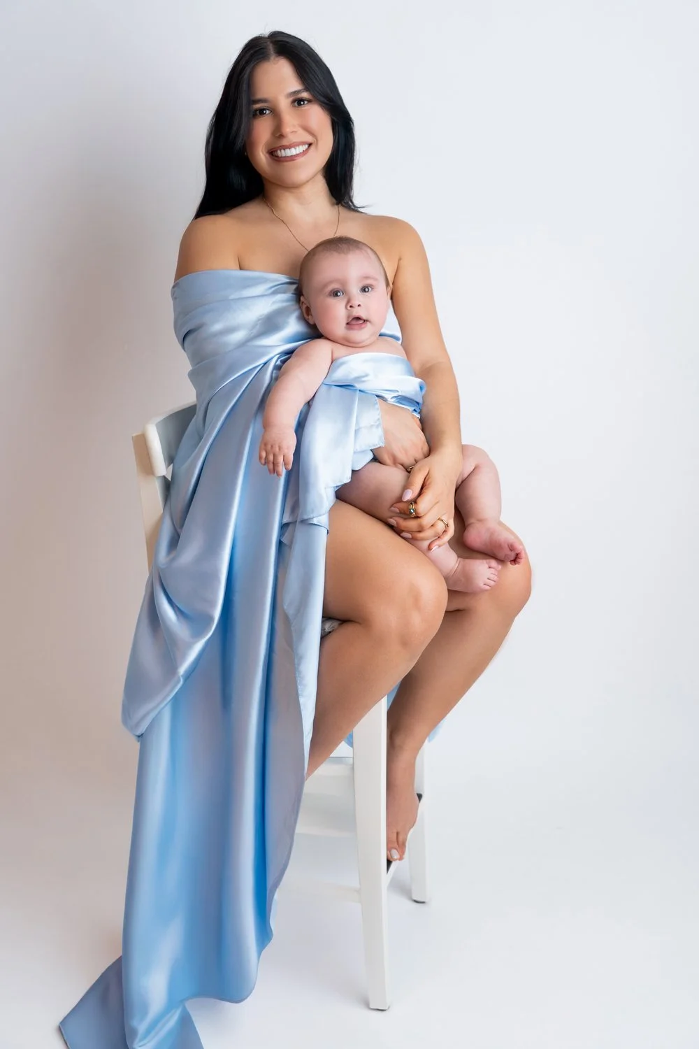 Frau mit schwarzen Haaren hält Baby in blauer Seidenschärpe auf einem weißen Stuhl vor einem weißen Hintergrund.