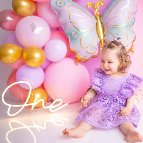 Mädchen im lila Kleid sitzt vor rosa Ballons und einer Schmetterlingskulisse.
