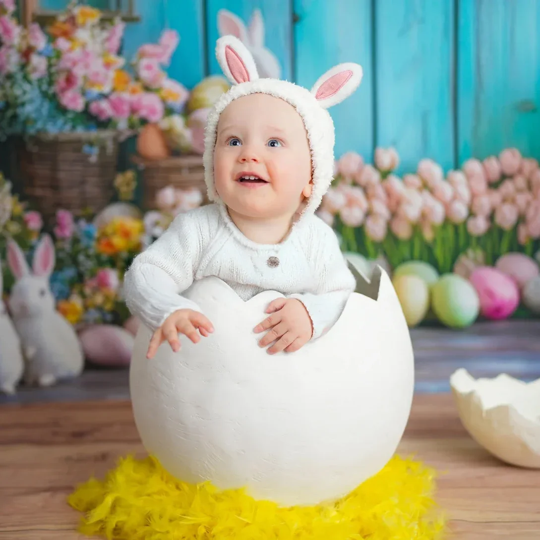 Ein Baby in einem Osterei-Kostüm mit Hasenohren, umgeben von Osterdekoration, Blumen und Osterhasen.