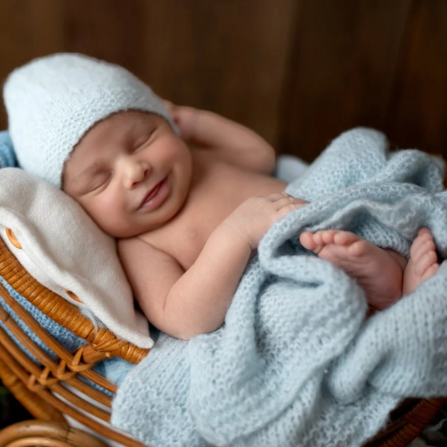 Ein schlafender Baby in einem Korb, eingewickelt in eine graue Decke, trägt einen weißen gestrickten Hut.