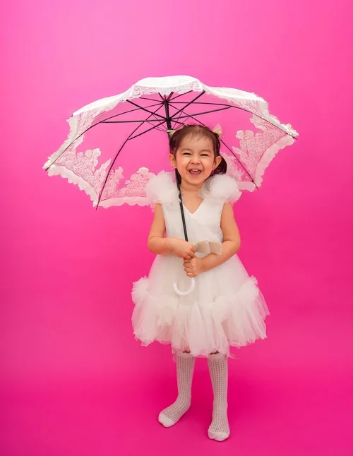 Lachendes Mädchen im weißen Kleid hält einen Schirm vor pinkem Hintergrund.