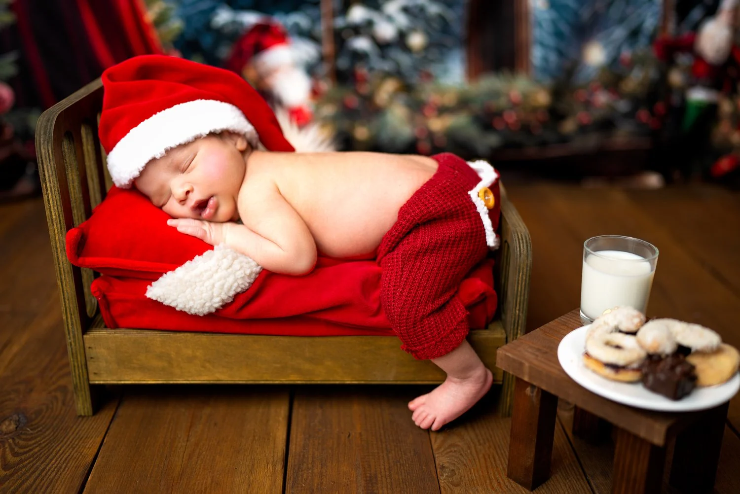 Ein schlafendes Baby in rot-weißer Weihnachtsmütze und Pullover liegt auf einem kleinen Bett mit rotem Kissen, umgeben von Weihnachtsdekoration.