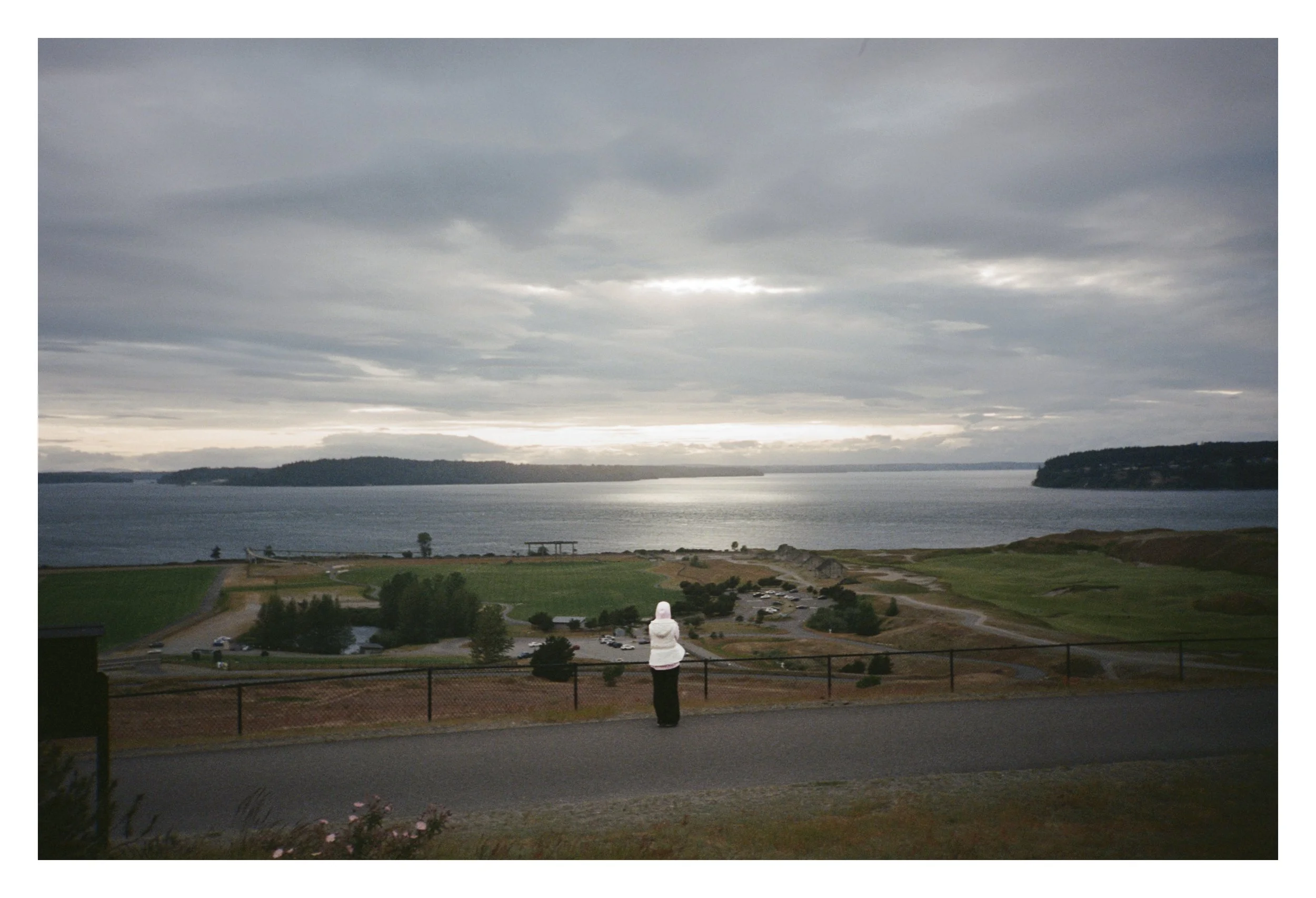 Person in white hoodie overlooking scenic view of the ocean, hills, and cloudy sky.