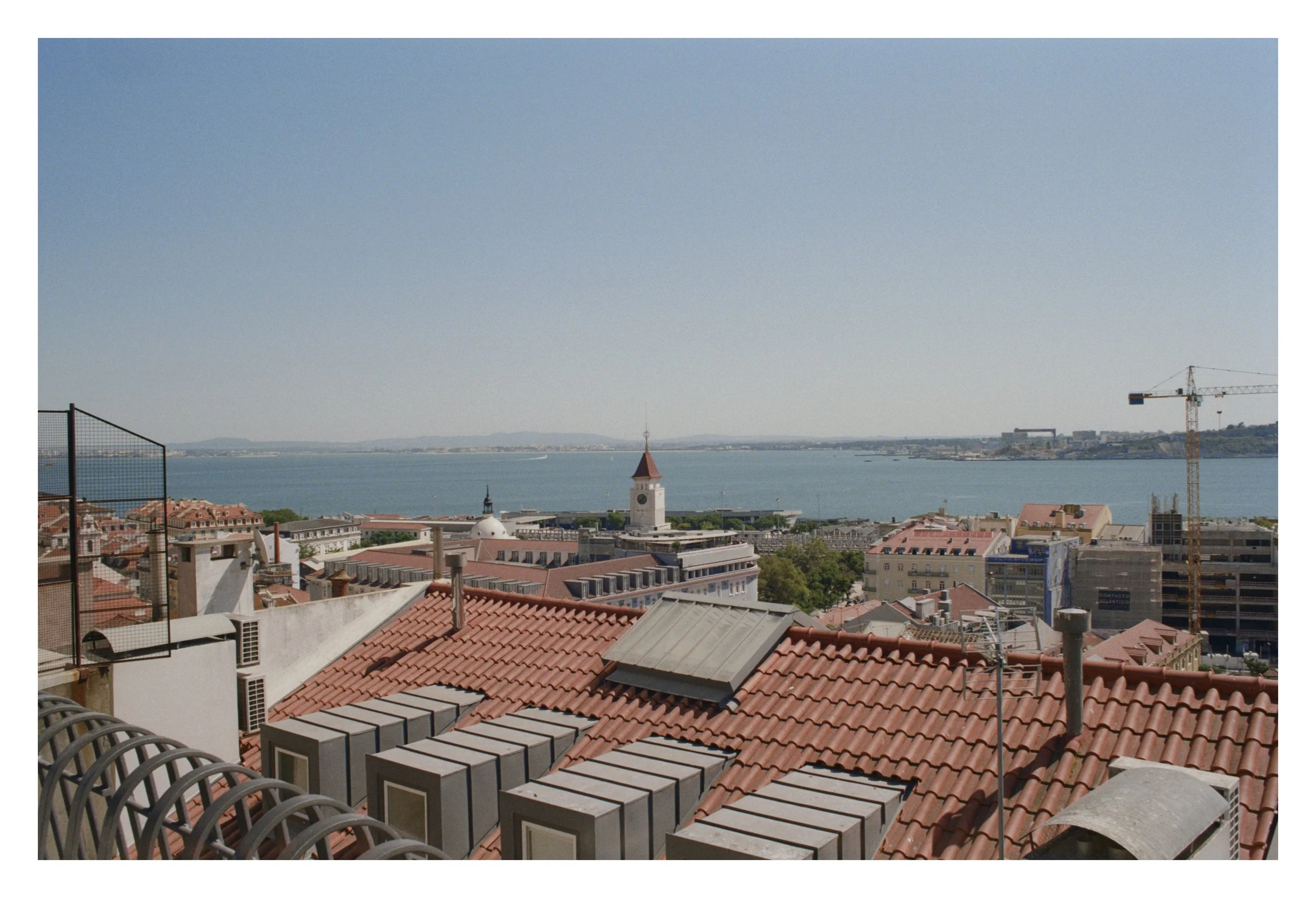 Cityscape with red tiled roofs, a distant body of water, and construction cranes under a clear blue sky.