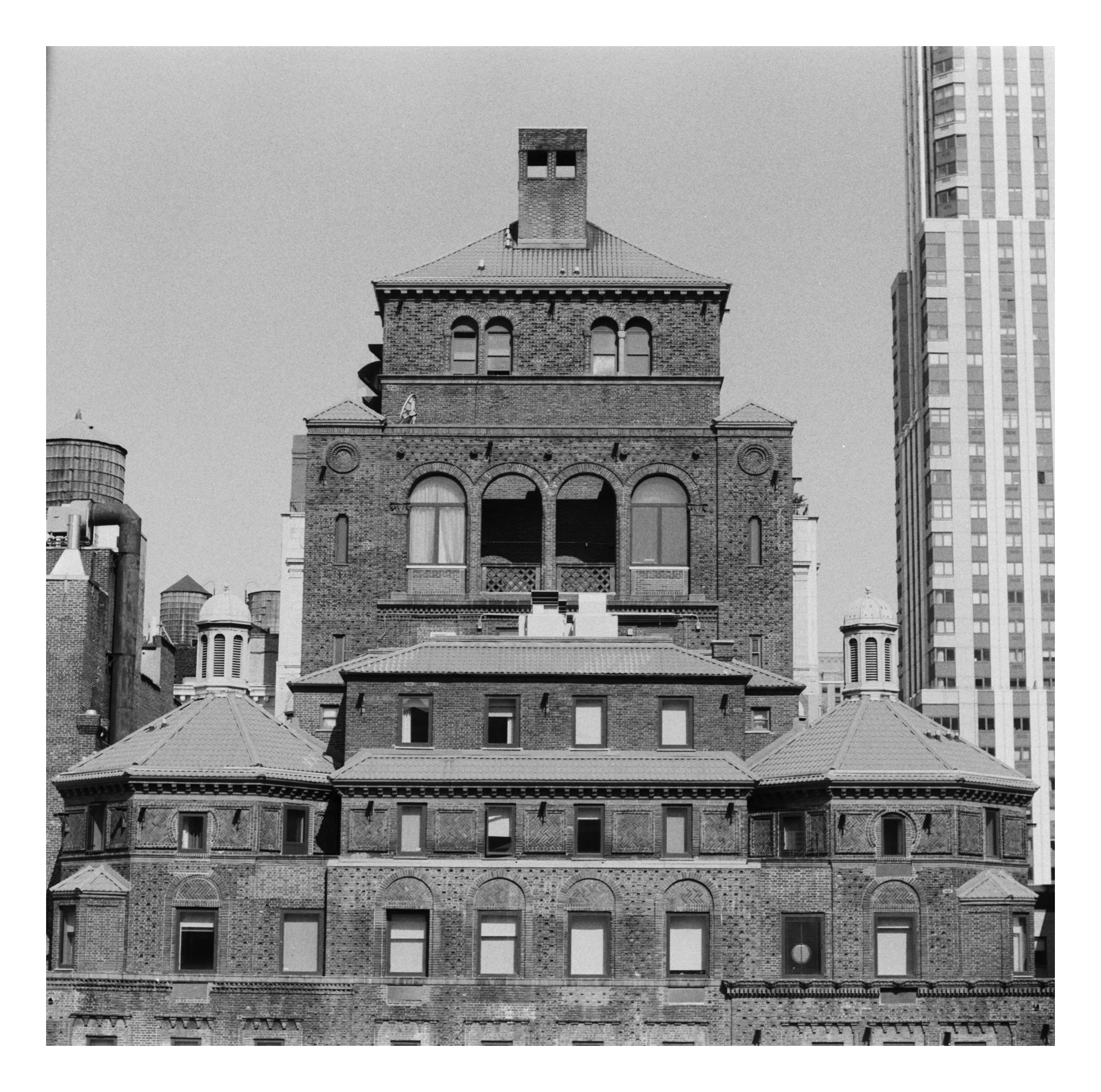 Black and white photo of a historic brick building with arched windows and decorative elements, surrounded by modern skyscrapers.