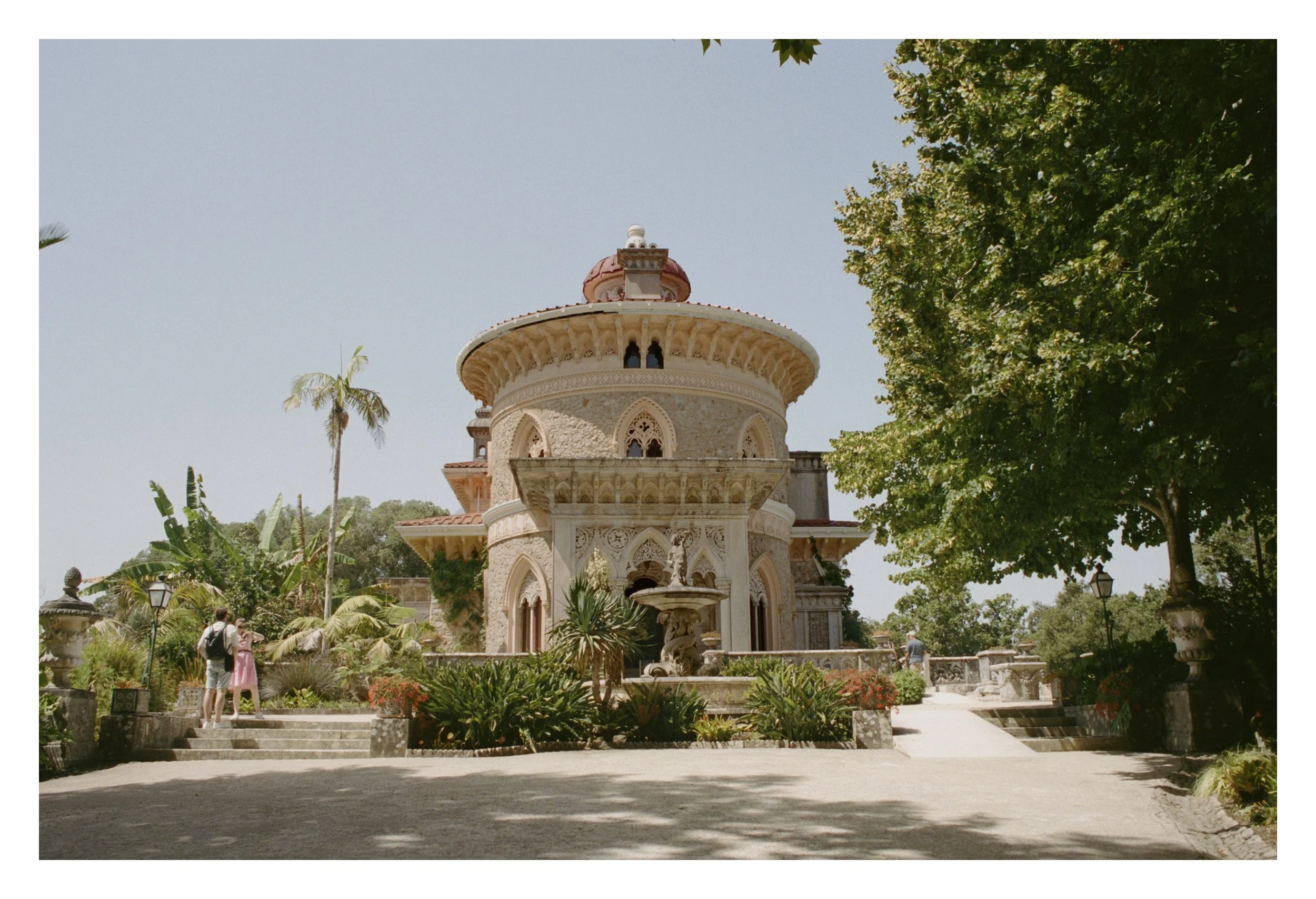 A historical building with intricate architecture surrounded by lush gardens and palm trees under a clear blue sky.