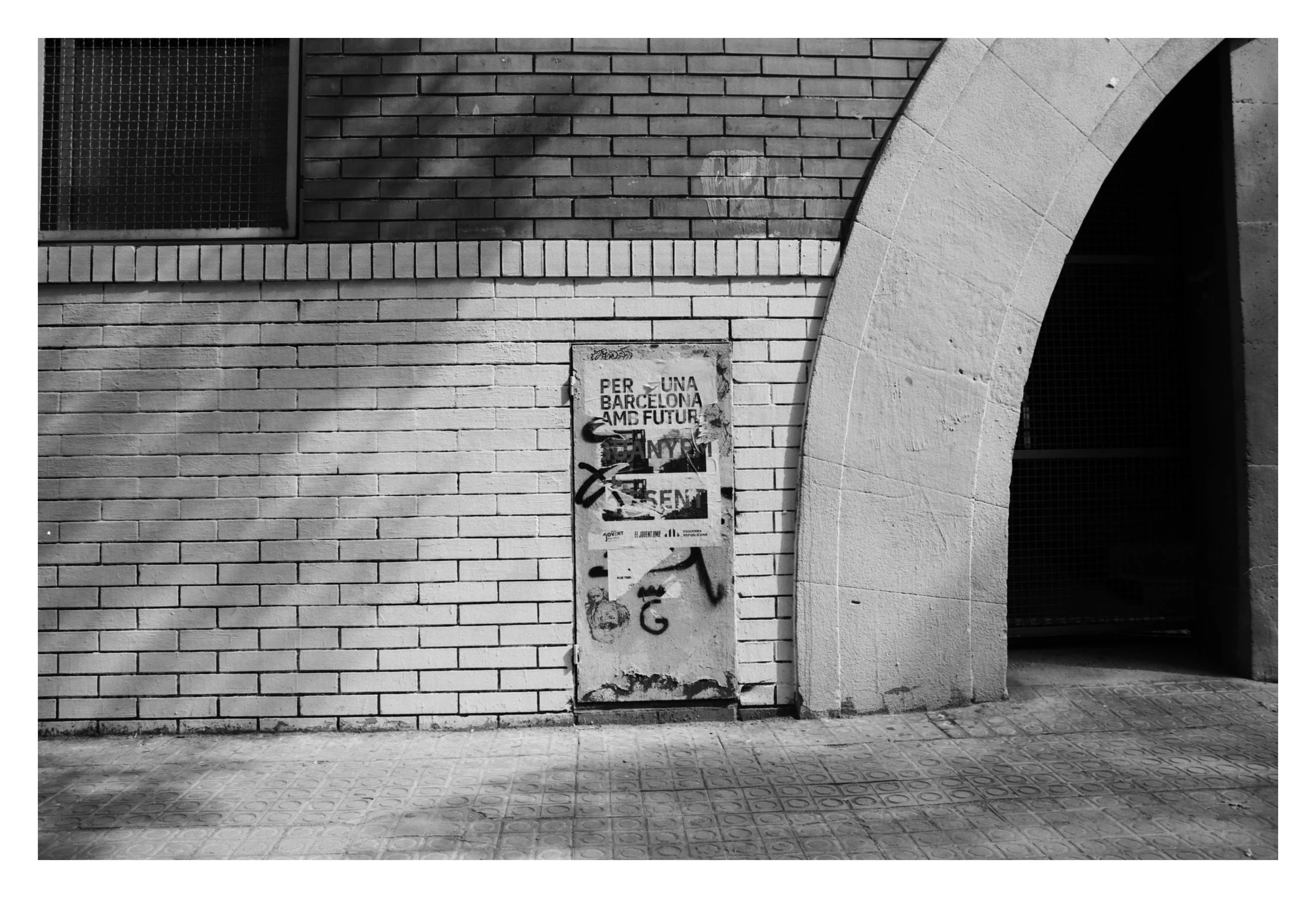 Black and white photo of a brick wall with a narrow door covered in graffiti and posters, next to an arched entrance.