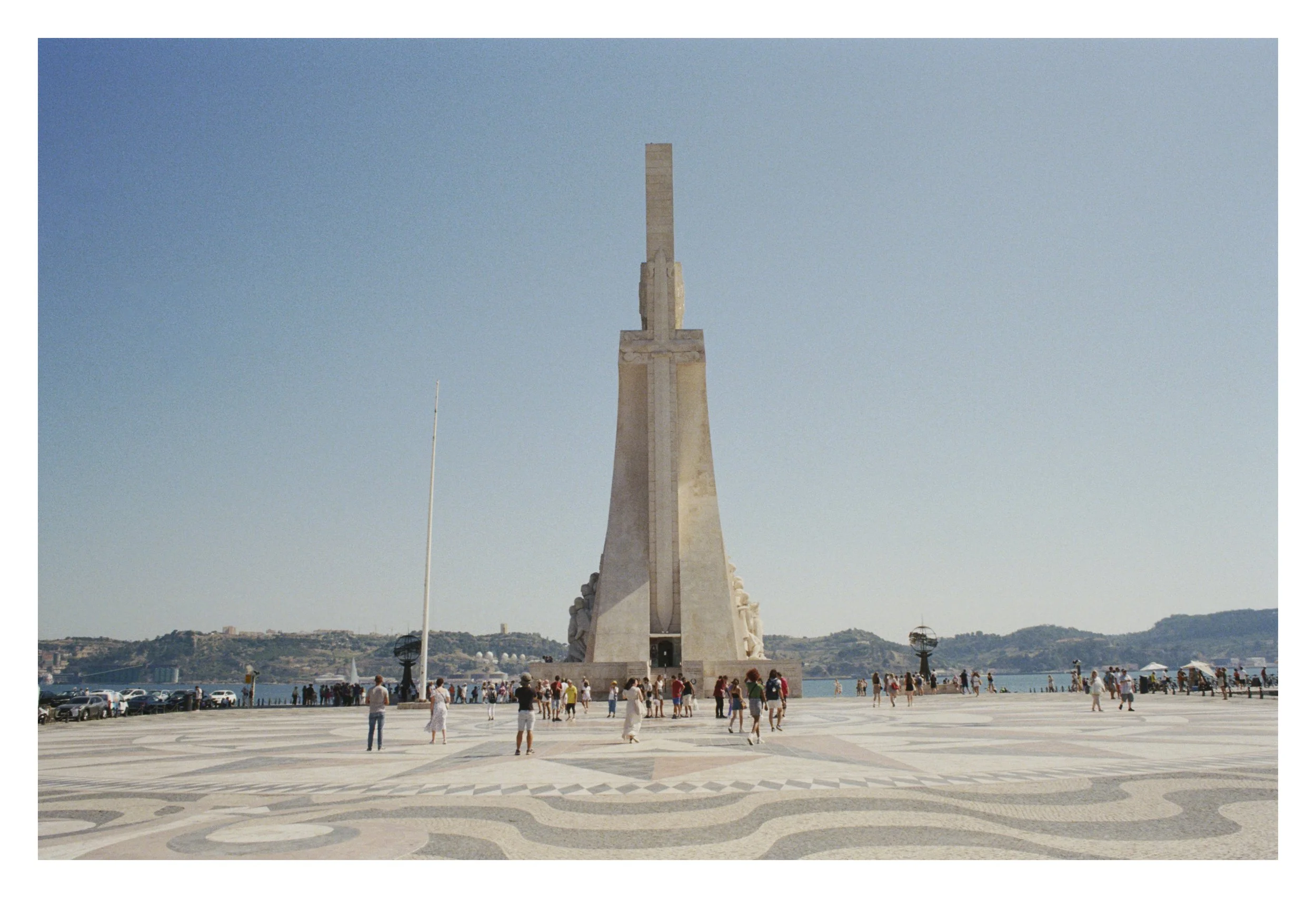 Monument with visitors, large stone structure, tiled plaza, sea in background, clear sky.
