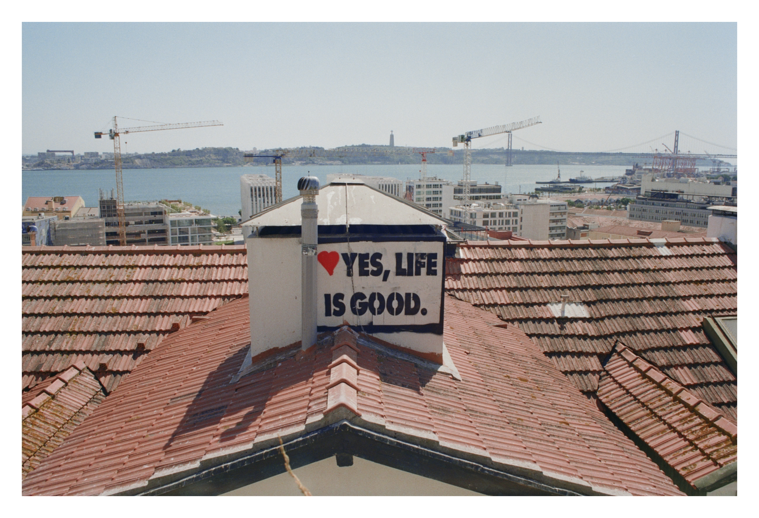 Tile rooftops with a sign reading '♥ YES, LIFE IS GOOD' in the foreground, overlooking a cityscape with a river, cranes, and a bridge in the background.