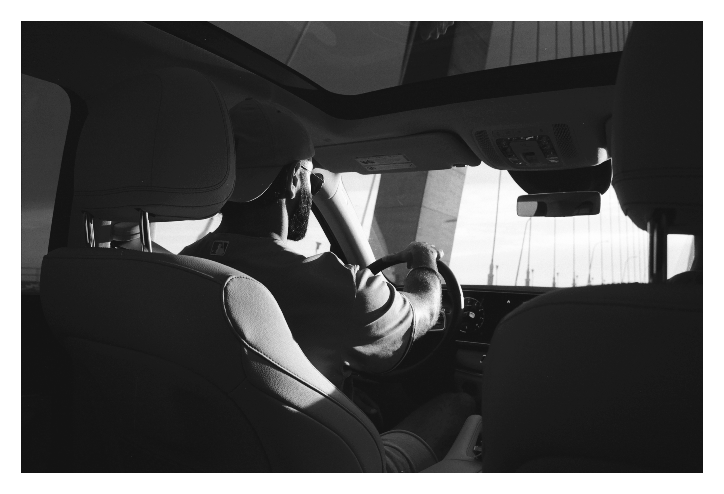 Back view of a person driving a car, wearing a cap and sunglasses, with interior car design visible and a bridge seen through the windows.