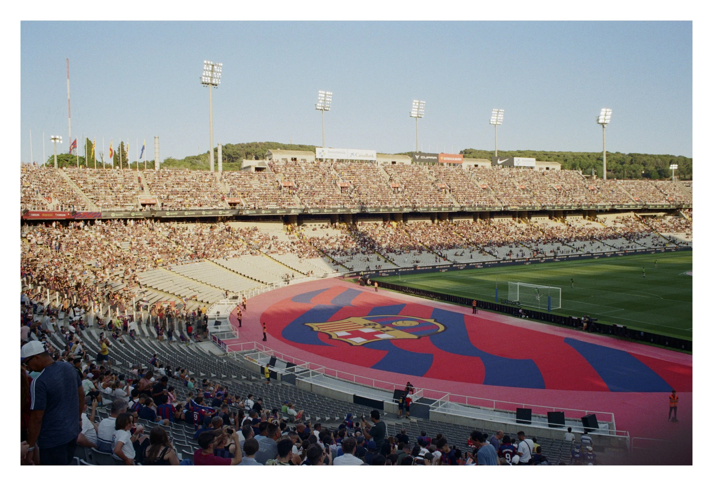Soccer stadium with audience, large FC Barcelona logo on field, and surrounding stands filled with spectators.
