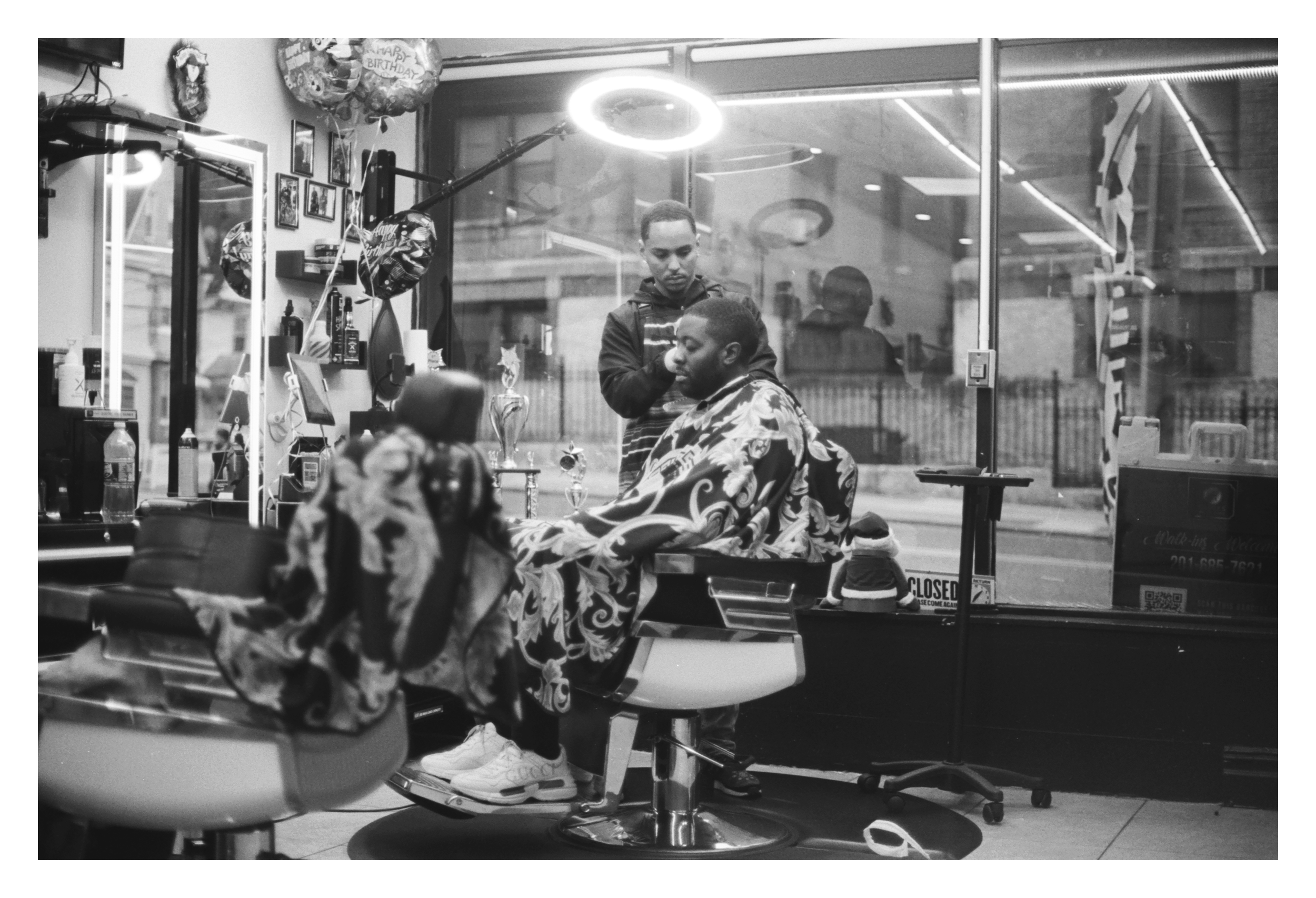 Black and white photo of a barbershop with a barber cutting a client's hair. The client is sitting in a barber chair covered with a patterned cape. The shop has a mirror, hair products, and decorations. A window displays a street view.