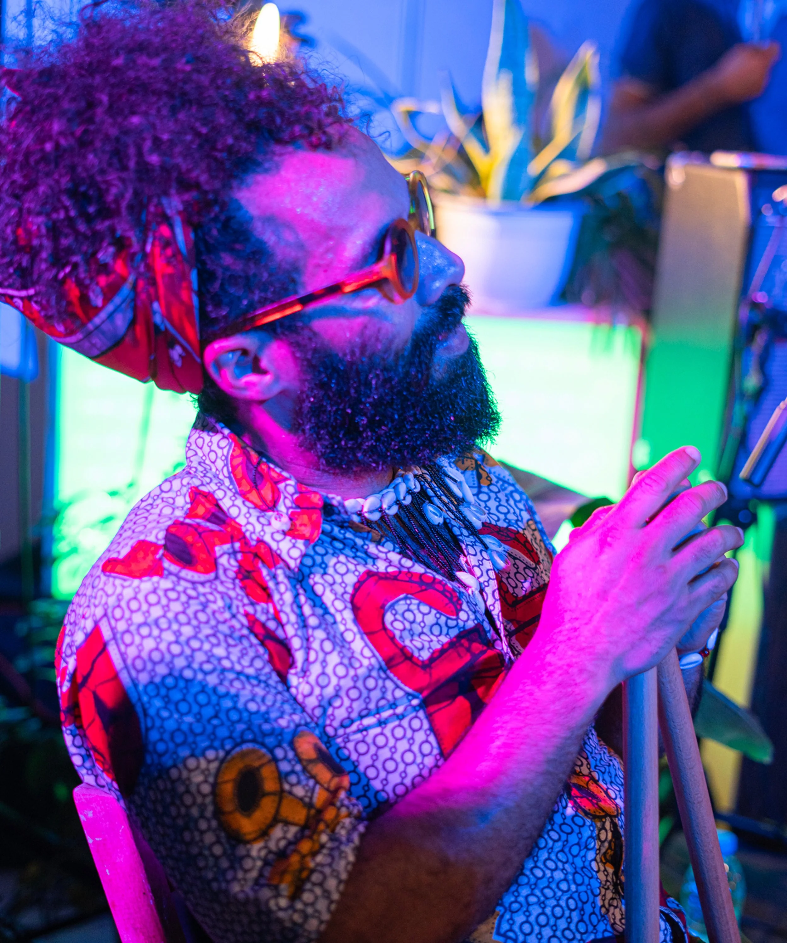 Persona con gafas de sol y cabello rizado, vestida con ropa de colores vivos, tocando un instrumento de percusión, con fondo de luces y plantas.