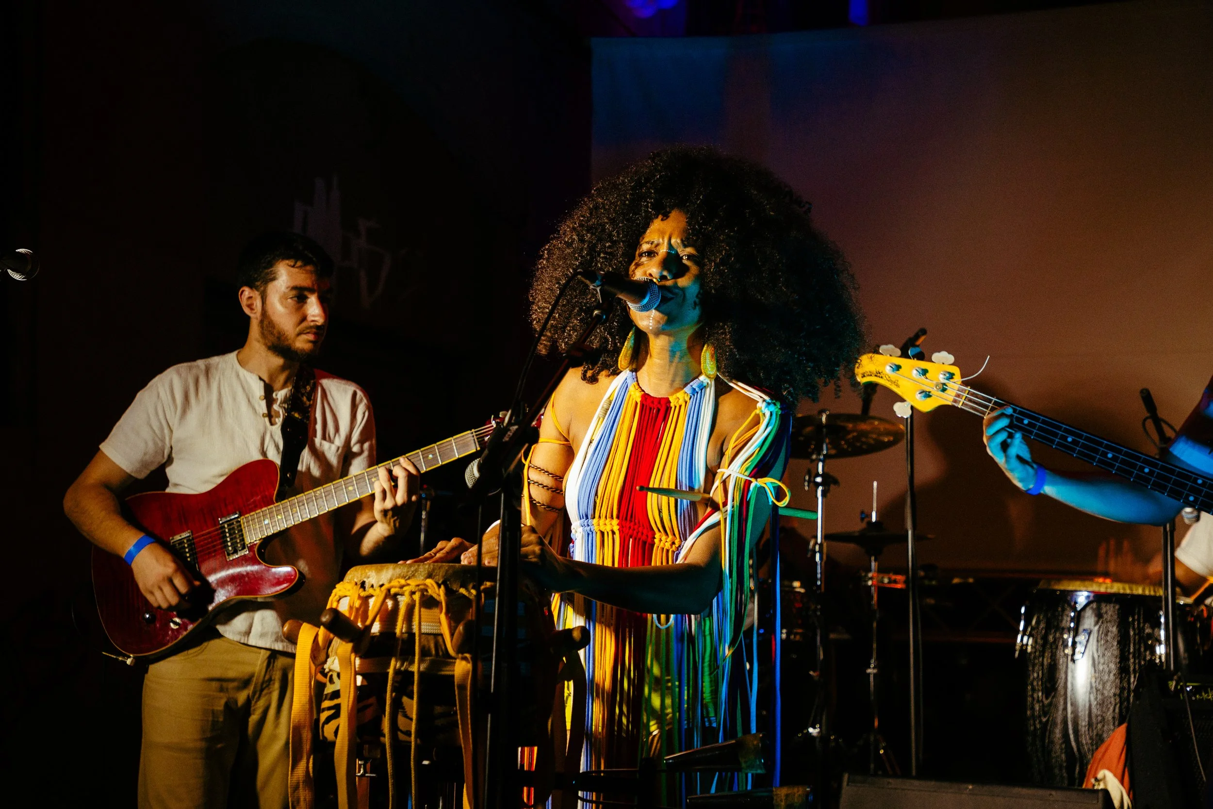 Una banda musical en vivo donde una mujer canta y toca instrumentos de percusión, acompañada por un guitarrista y un bajista. La iluminación es tenue y colorida.