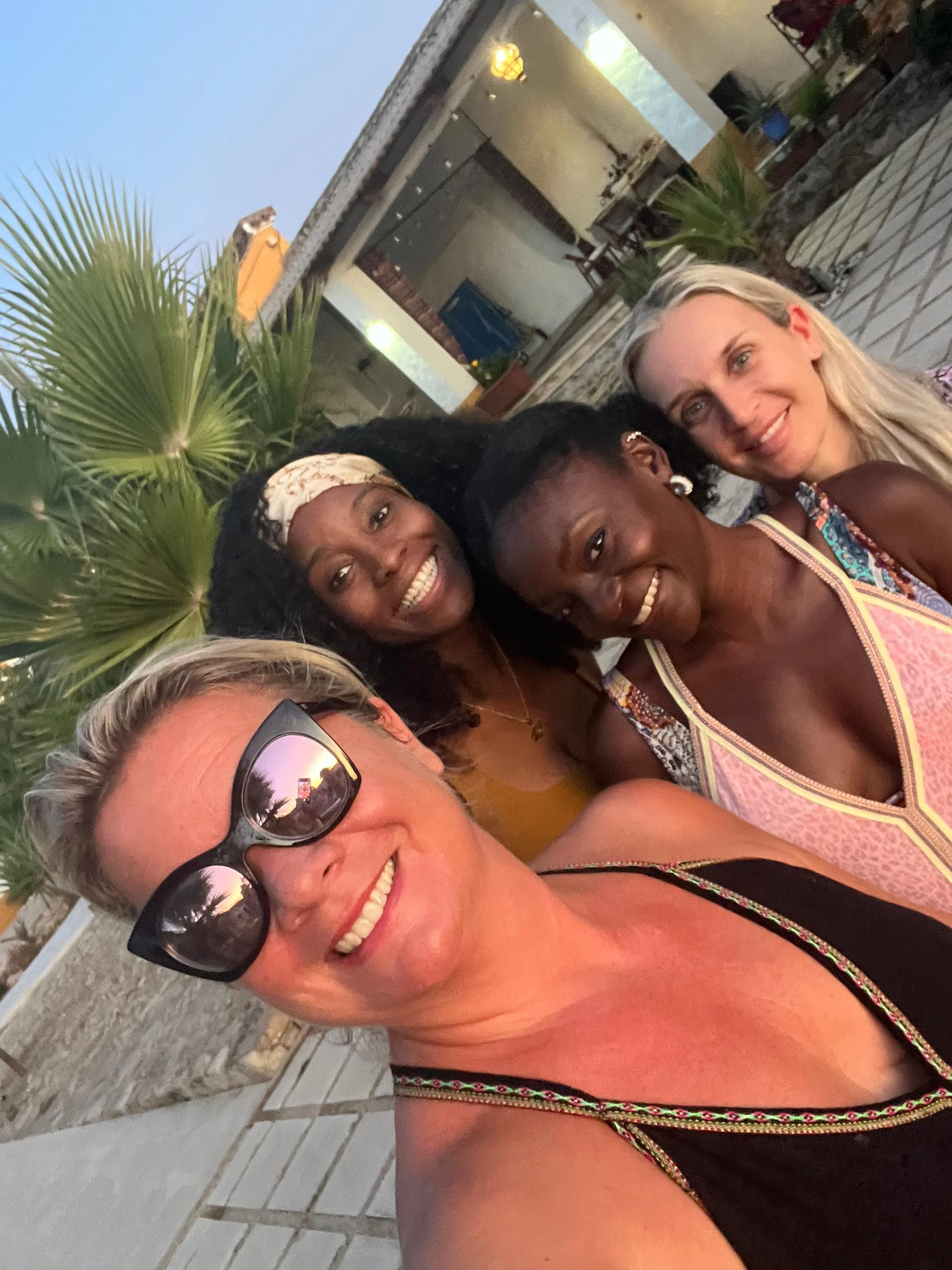 View through a dark wooden window frame showing an outdoor scene with lemon trees, a patio with chairs, and a white building under a partly cloudy blue sky.