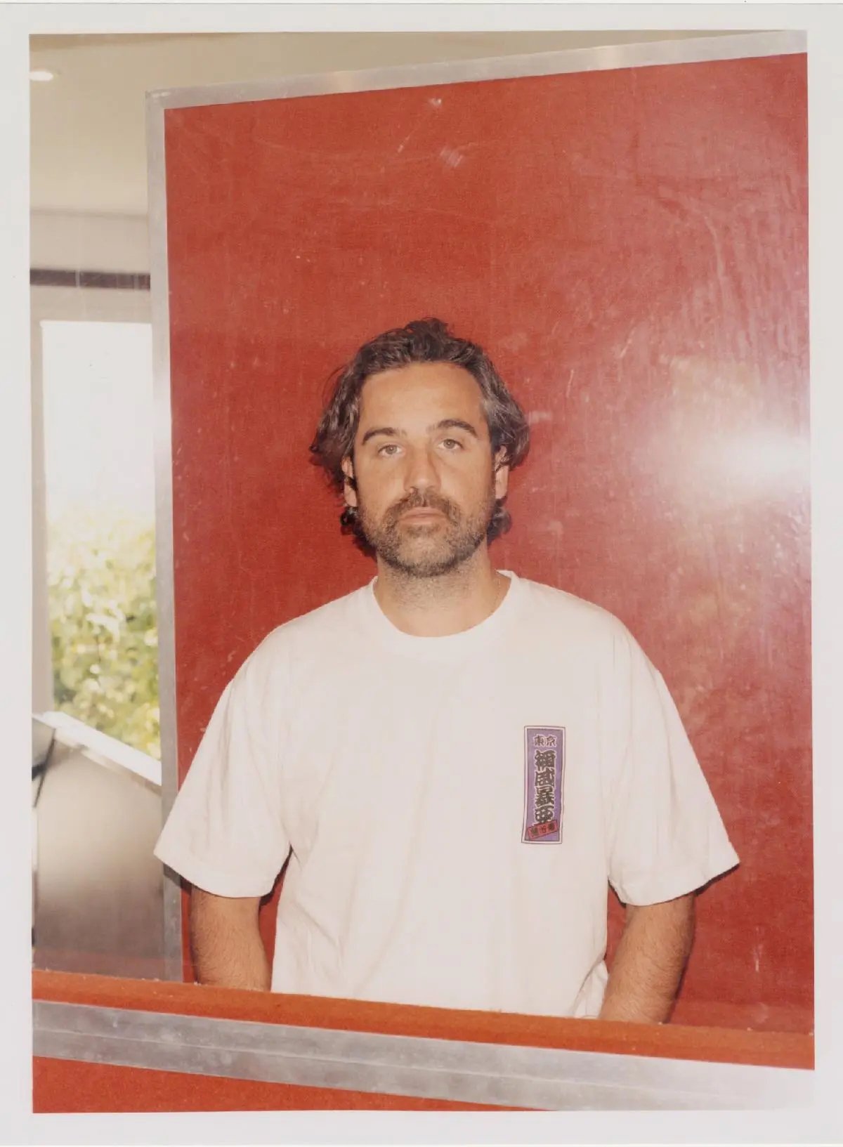 A person with medium-length hair and a beard, wearing a white t-shirt, standing against a red background with a reflective surface.