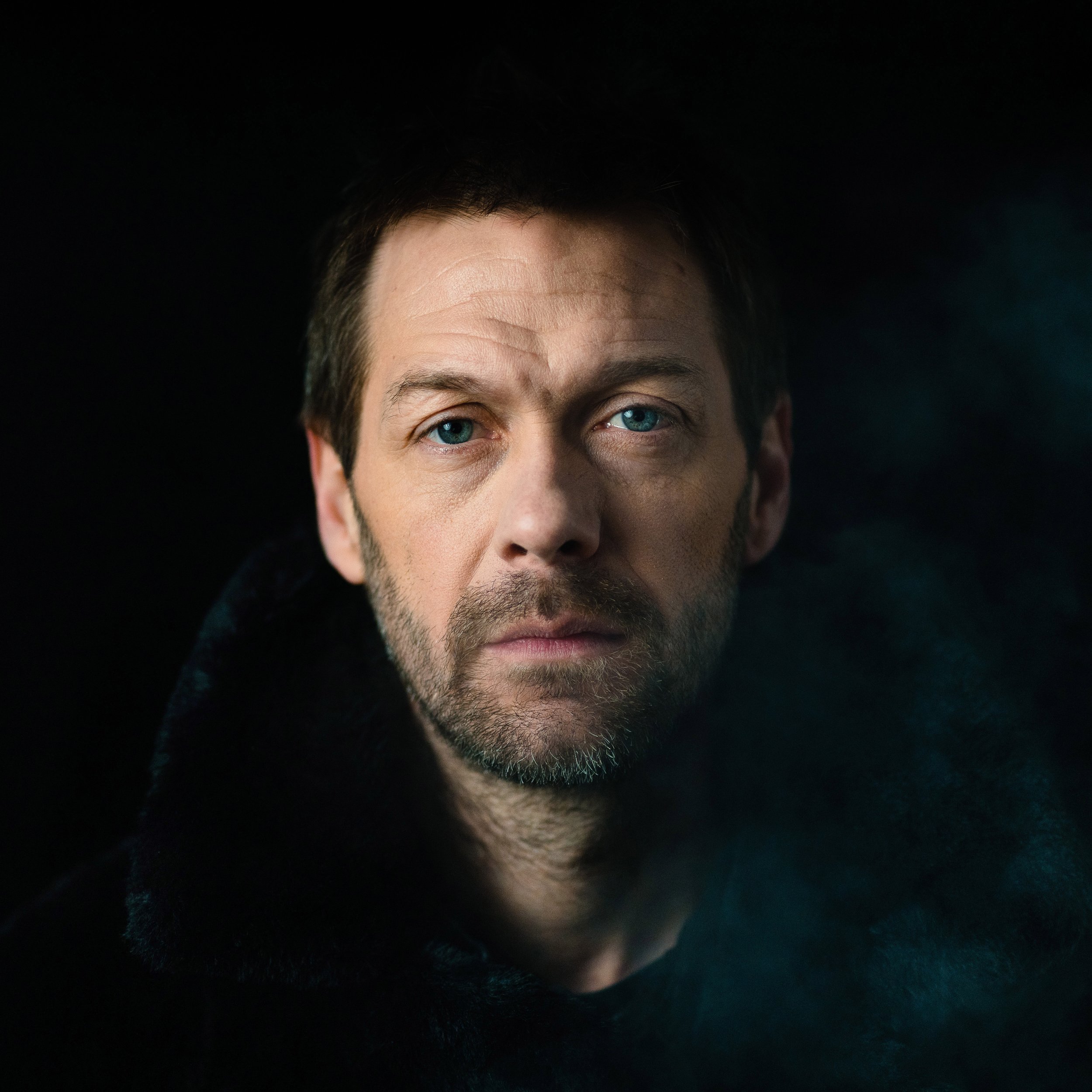 Portrait of a man with blue eyes and a beard wearing a dark coat against a black background.