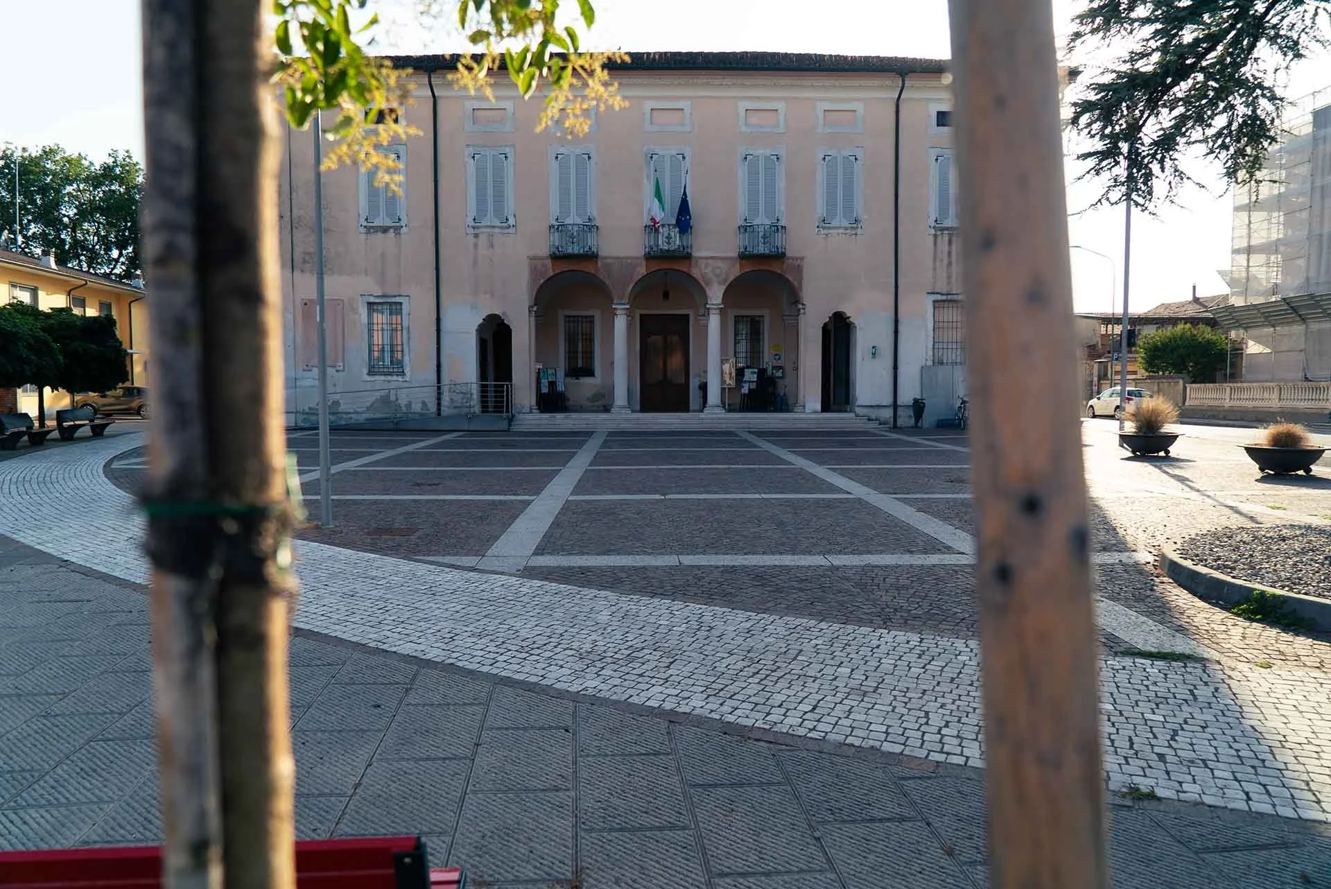 Edificio pubblico in stile classico con bandiere italiane ed europee, vista da dietro una ringhiera, in una piazza con pavimentazione in pietra, panchine e piante ornamentali.