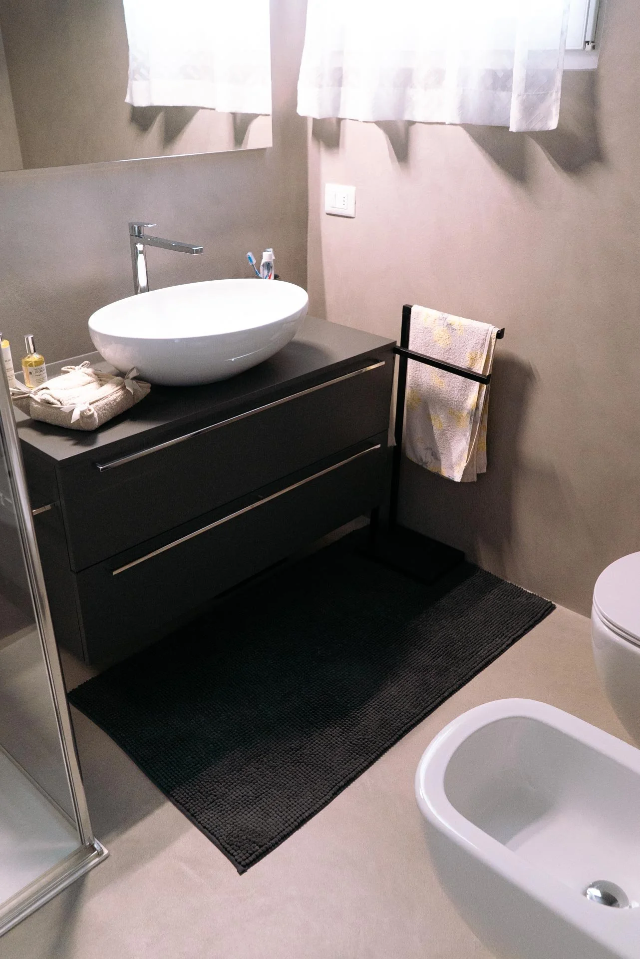 Bagno moderno con lavabo su mobile scuro, specchio, asciugamano appeso, bidet bianco, e tende leggere chiuse sulla finestra.