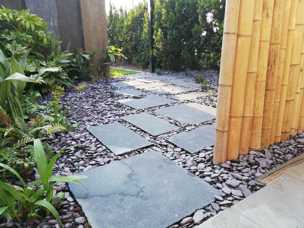 Pavimentazione con piastrelle di pietra grigia lungo un passaggio da giardino, con vegetazione verde e pareti in legno e muri laterali.