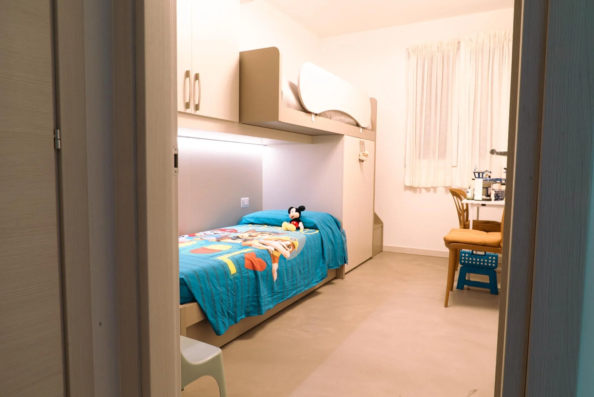 Stanza per bambini con letto singolo, coperte colorate con personaggi dei cartoni, bambola di Mickey Mouse sopra il letto, scrivania con oggetti scientifici, sedia imbottita e finestra con tende leggere.