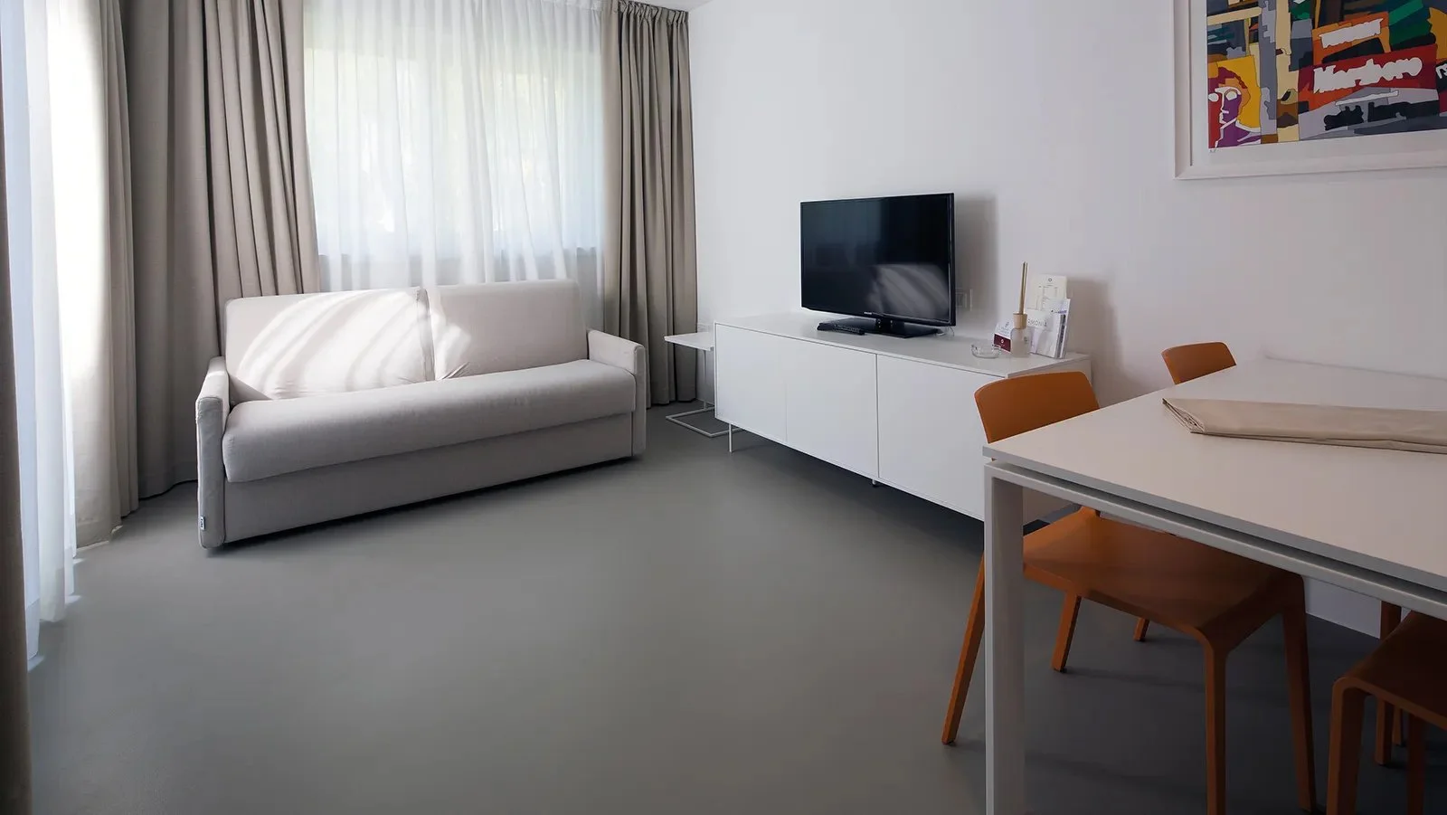 Interno di un soggiorno elegante con divano bianco, televisore su mobile bianco, tavolo con sedie marroni, pareti bianche e dipinto colorato.