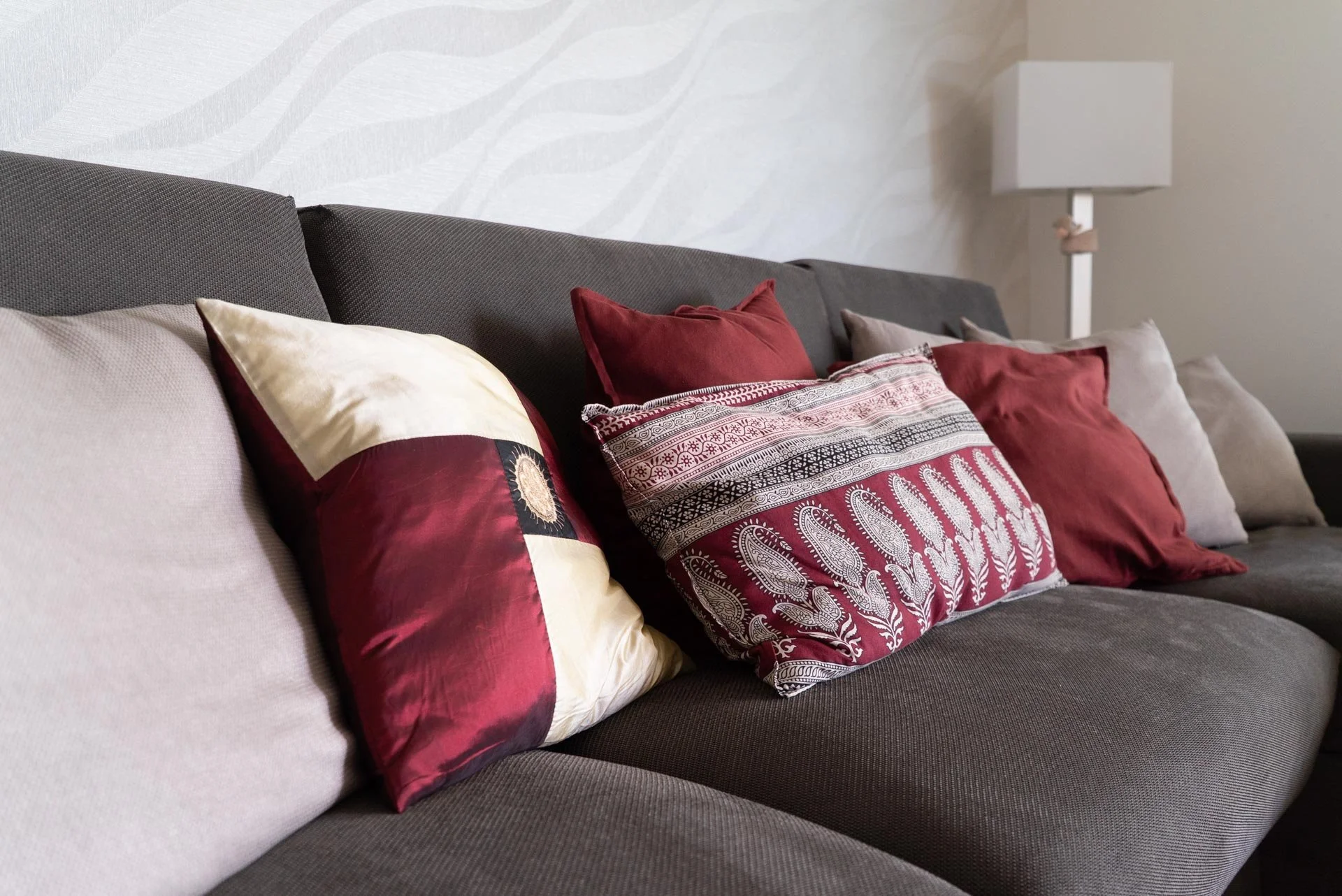 Cuscini di vari colori e fantasie su un divano grigio in un soggiorno con parete chiara e lampada da terra.