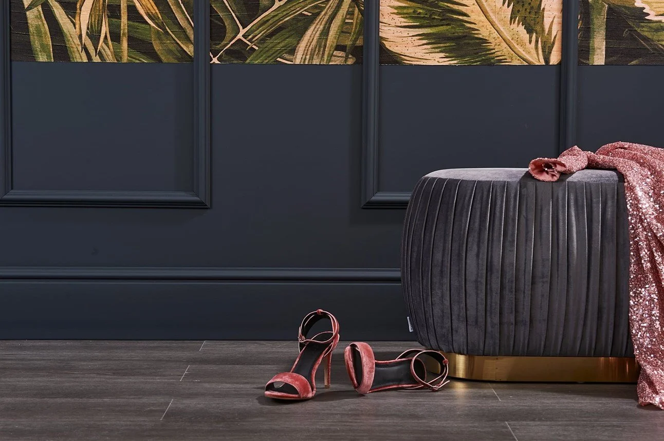 Un paio di scarpe rosse con tacco alto e una sciarpa di paillettes rosa sono posate su un sedile imbottito di velluto grigio scuro, in una stanza con pareti blu scuro e decorazioni con pannelli con foglie tropicali.