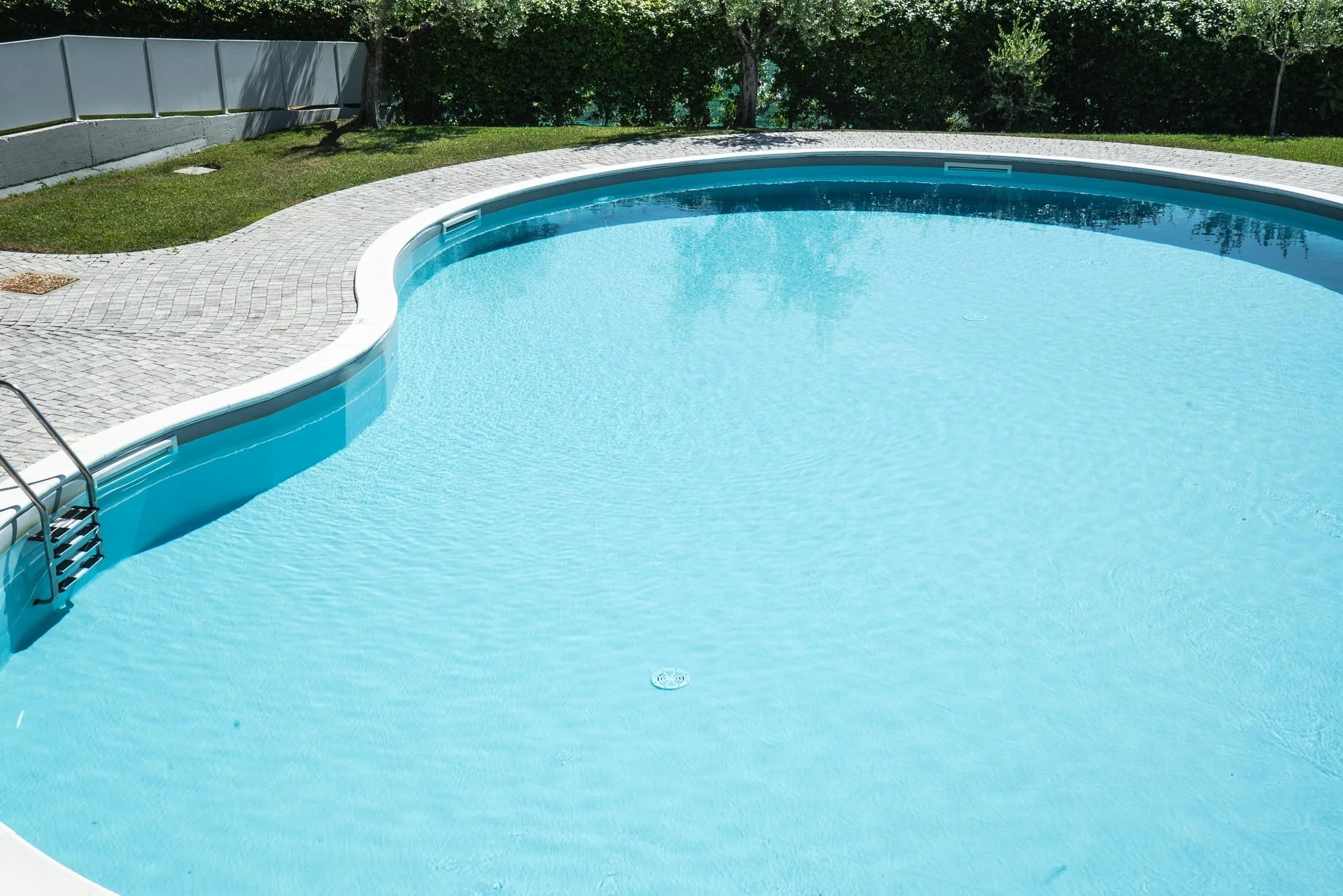 Piscina all'aperto circondata da pavimentazione in pietra, con alberi e siepi sullo sfondo.