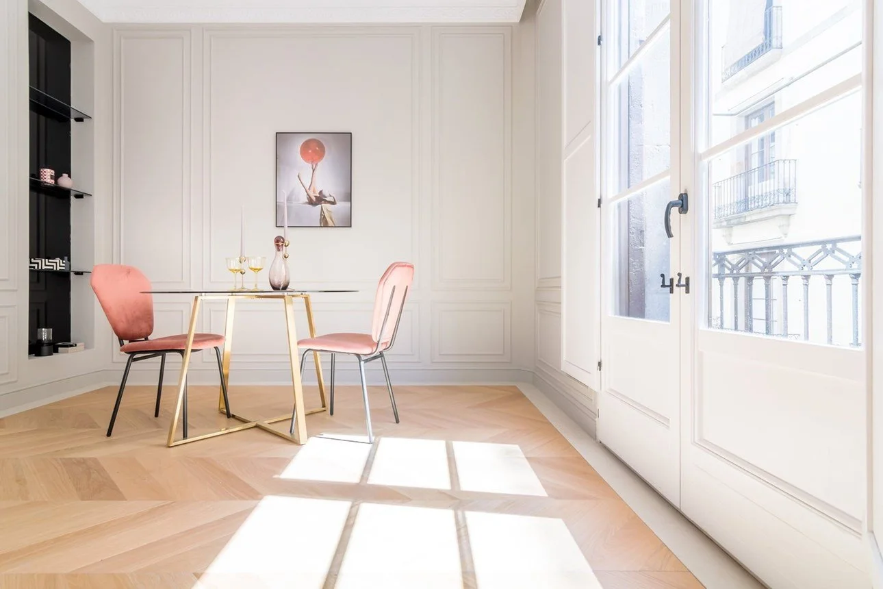 Sala da pranzo luminosa con due sedie rosa e un tavolo con candelabri, pareti bianche con pannelli decorativi, finestra grande che permette ingresso di molta luce.