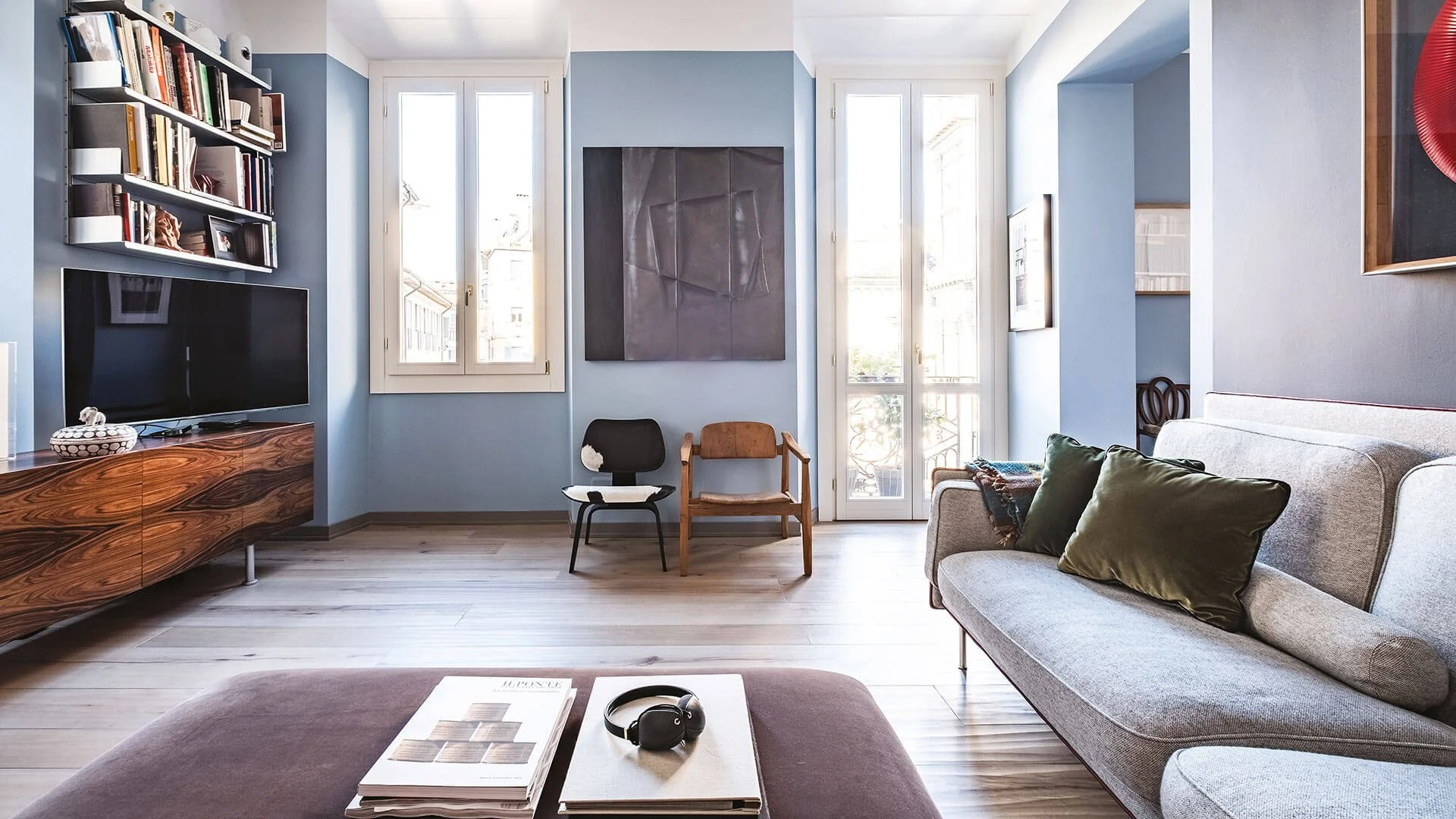 Soggiorno con divano grigio, due sedie di legno e una tv su mobile in legno, pareti azzurre con quadri e libreria. Finestre luminose con vista sui edifici.