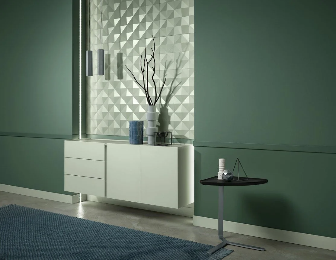 Un muro verde con una nicchia decorativa in bianco e marrone chiaro, con oggetti d'arredo e vasi con rami. Un tavolino nero con oggetti decorativi sulla destra e un tappeto blu sulla parte inferiore.