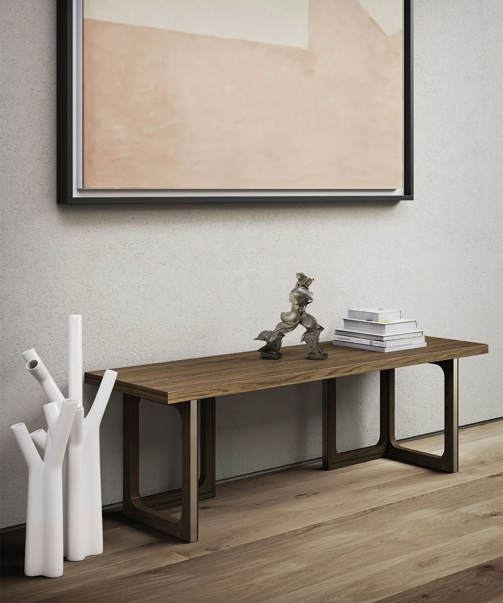 Scaffale basso in legno con cavalletto in metallo, decorazioni in metallo astratte e una pila di libri bianchi con copertina rigida, ai piedi un vaso decorativo bianco di forma simile a un albero privo di foglie, sullo sfondo una parete di colore chi