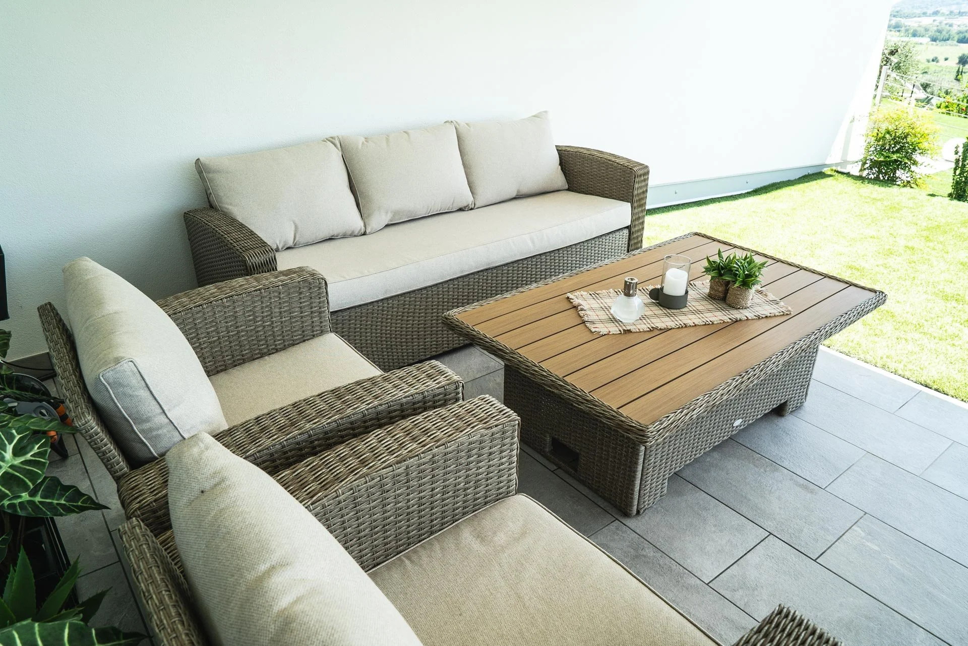 Arredamento da terrazza con divani in rattan, tavolino con pianta, candela, e ambiente esterno con vista su giardino verde.