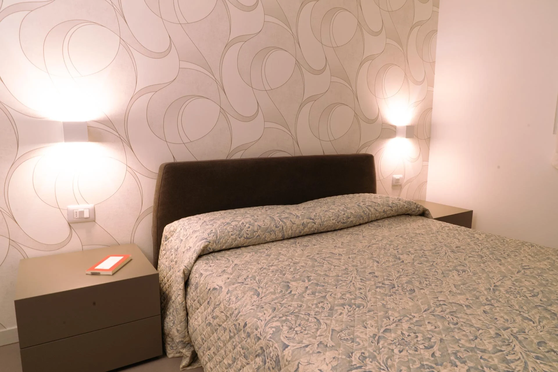 Letto con testiera neutra in una stanza con pareti decorate con motivo circolare, luci notturne ai lati del letto e un comodino con un block notes sopra.