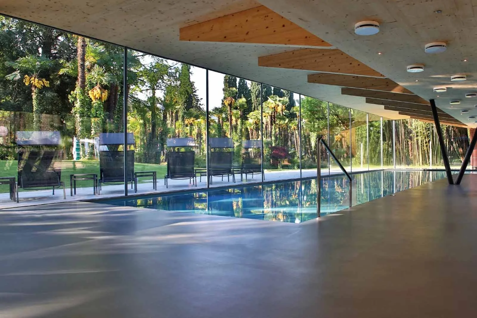 Piscina coperta con pareti di vetro che mostrano un giardino con alberi e sedie lounge.