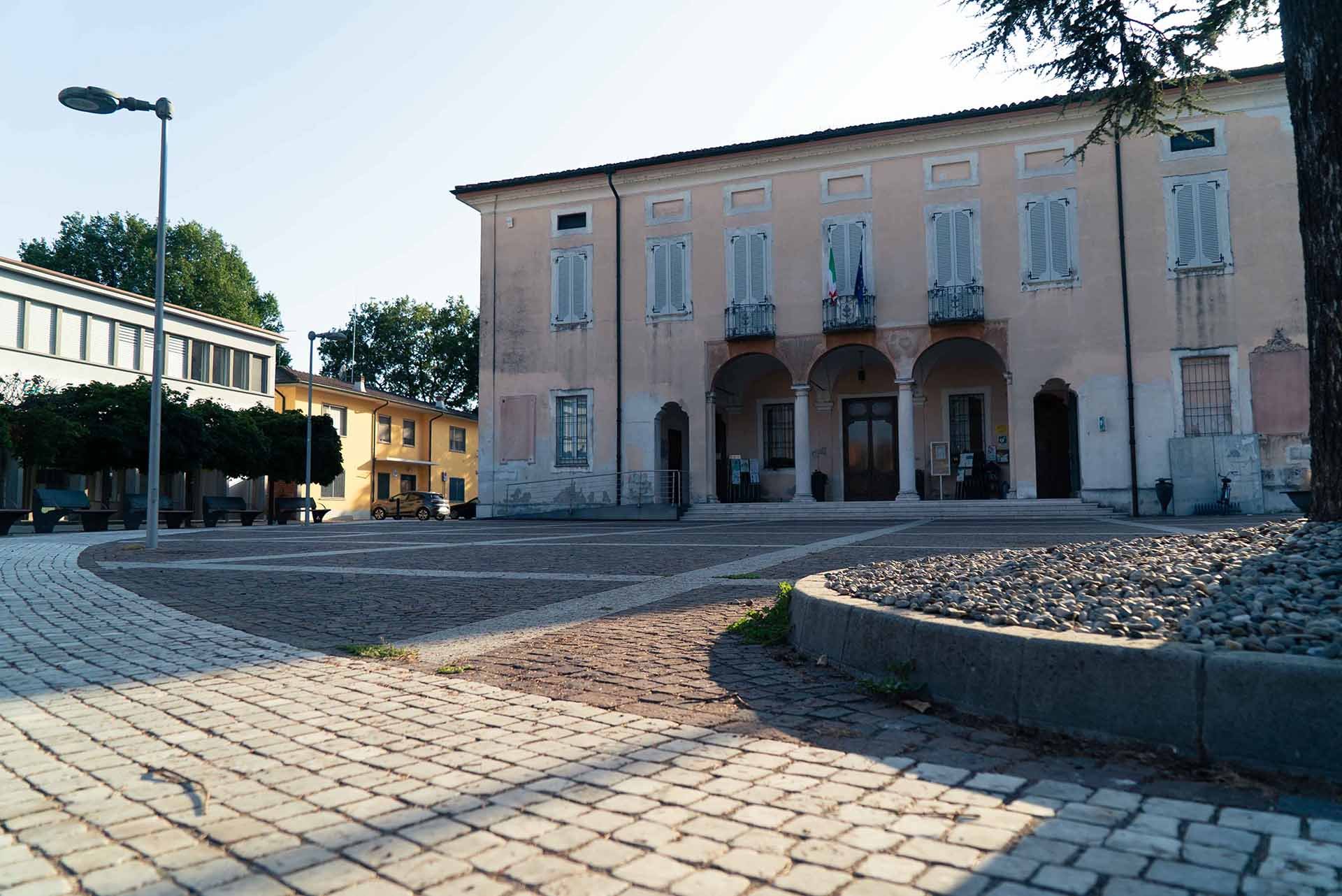 Piazza con edificio storico in stile italiano, con tre archi e bandiere, sedie e panchine, lampioni e panchine, pavimentazione in pietra, alberi e strada tranquille.