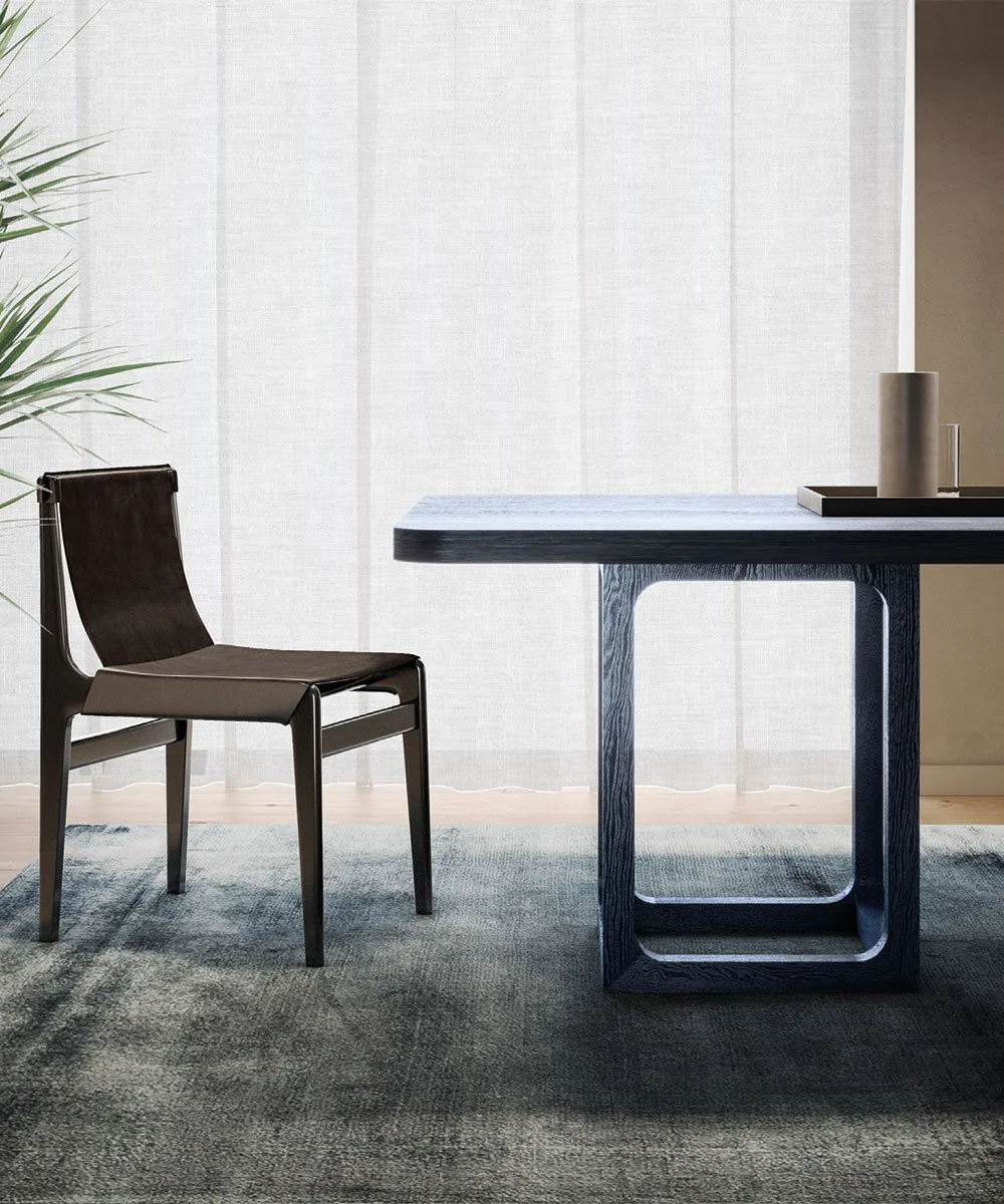 Interno con tavolo in legno scuro, sedia moderna in pelle e metallo, tappeto grigio e tende chiare.