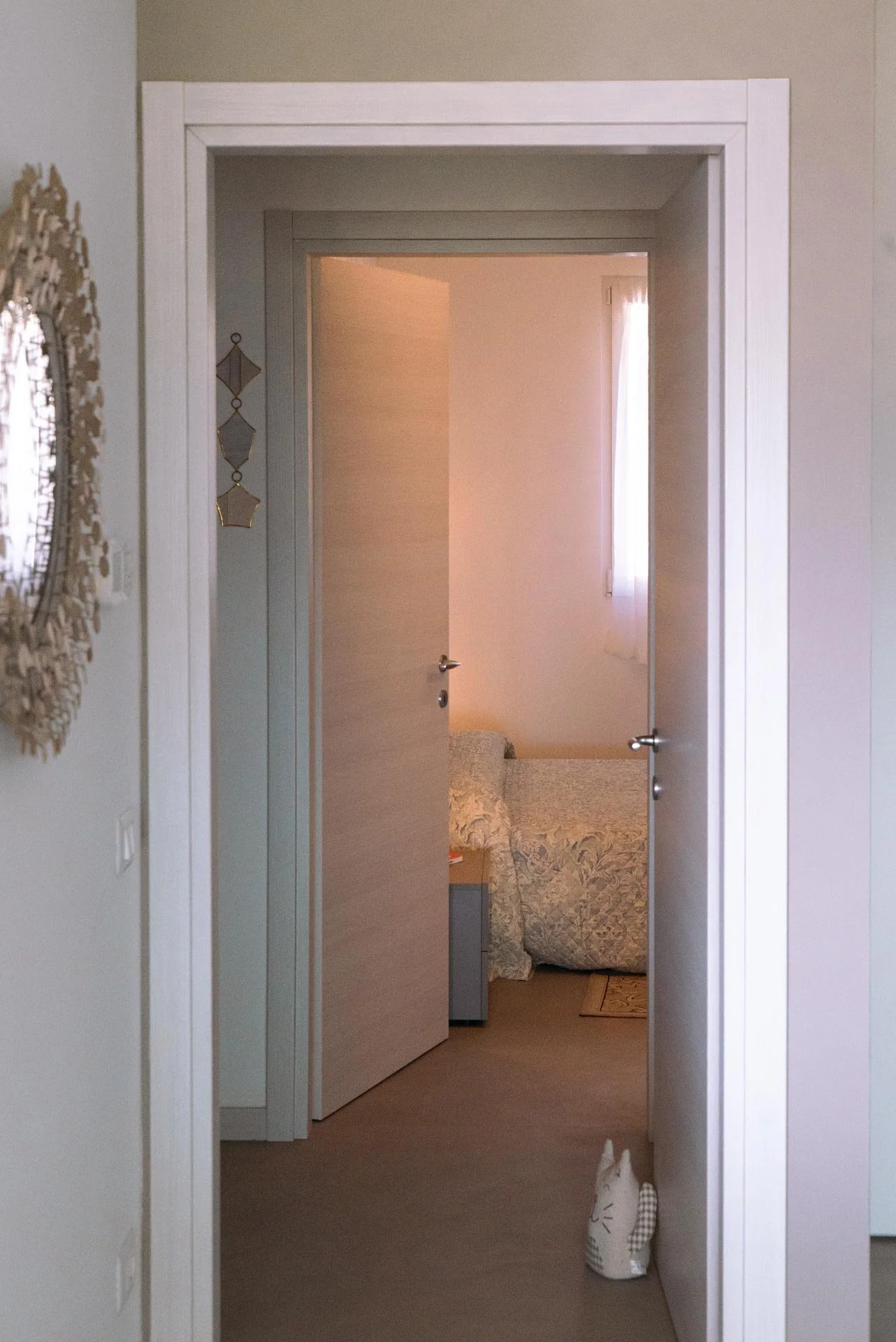 Vista di un corridoio con una porta aperta che conduce a una camera da letto con un letto e una finestra con tende leggere. L'interno è arredato in stile semplice e minimalista, con pareti chiare e decorazioni morbide.