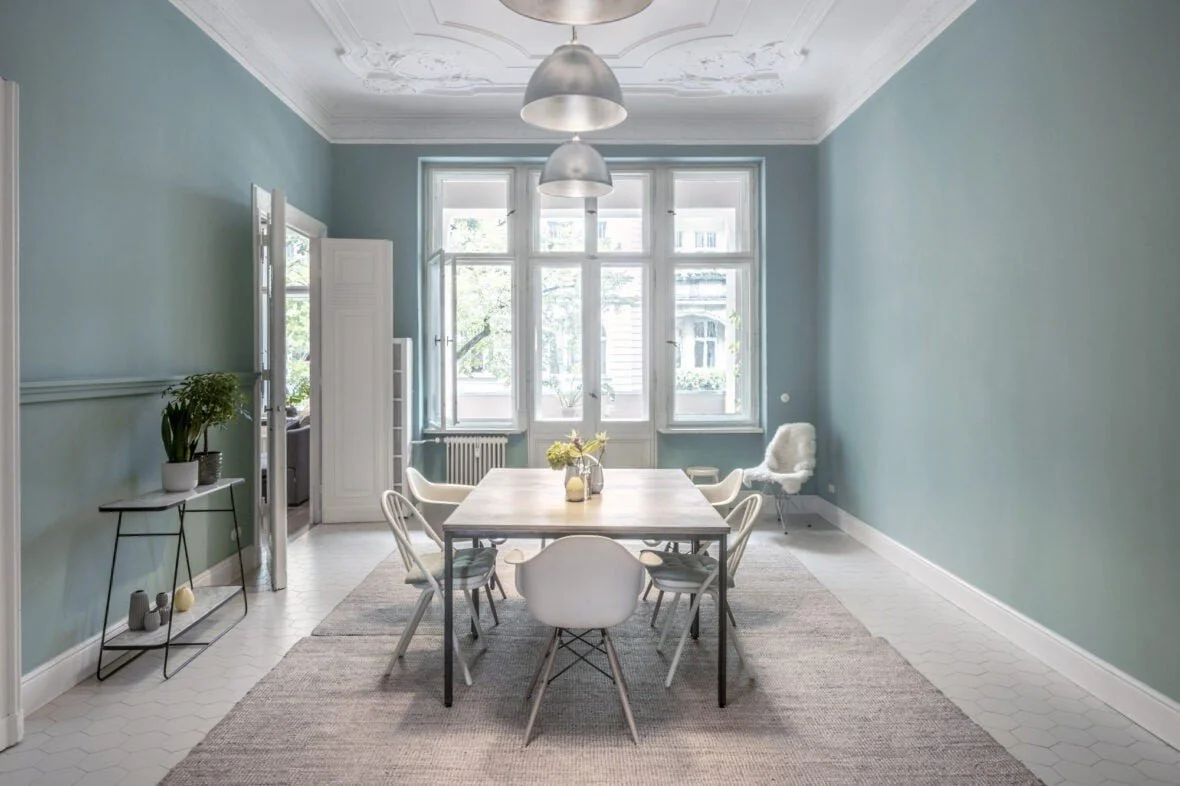 Sala da pranzo luminosa con pareti verdi acqua, grande finestra, tavolo in legno circondato da sedie bianche, lampade metallizzate al soffitto, e decorazioni minimaliste