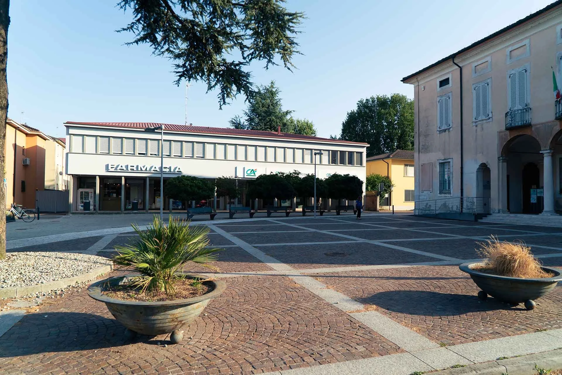 Piazza con aiuole e panchine, edificio commerciale con farmacia, e una chiesa con entrata e scalini.