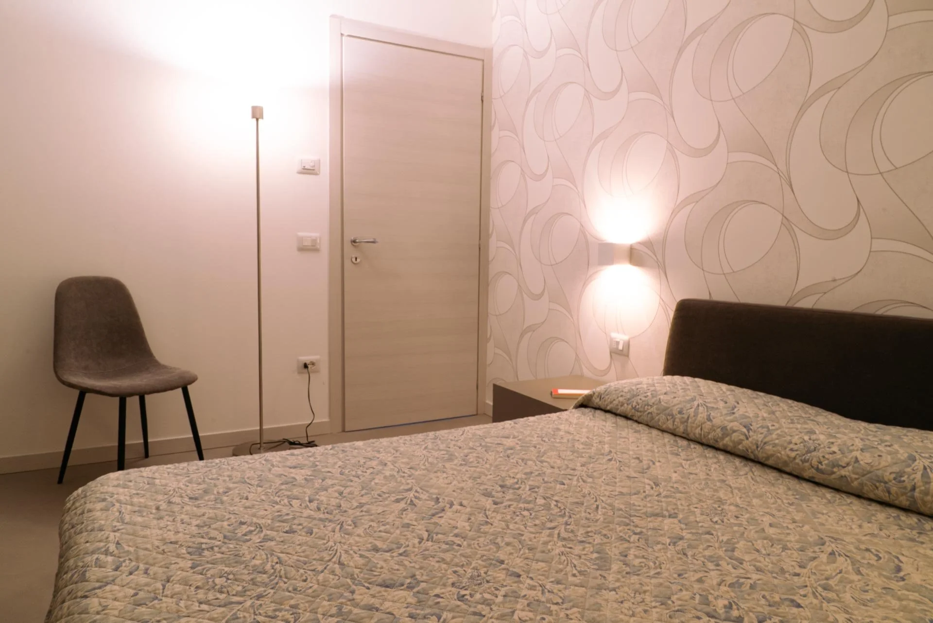 Camera da letto con letto matrimoniale, comò, sedia, lampada da terra, illuminazione soffusa e pareti decorate con disegni astratti.