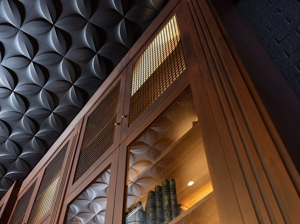 Particolare di un mobile in legno con ante in vetro e una libreria interna con libri antichi, sul soffitto un pannello 3D nero con motivi geometrici