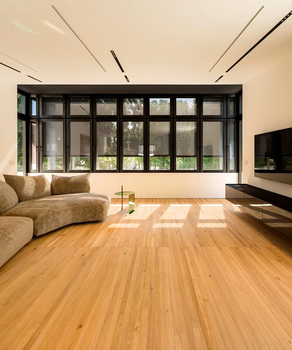 Interno di soggiorno moderno con grandi finestrone, divano beige, tavolino verde, pavimento in legno, televisore a parete e pareti bianche.