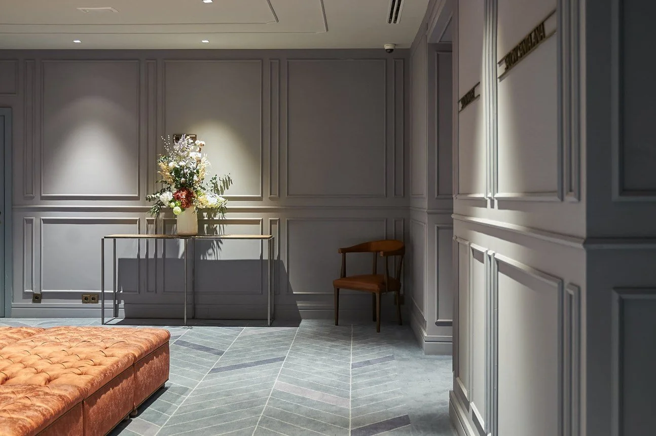 Interno di una stanza elegante con pareti rivestite di pannelli, un tavolo con un grande vaso di fiori e un divano in velluto rosa. C'è anche una sedia di legno e un pavimento con un motivo geometrico.