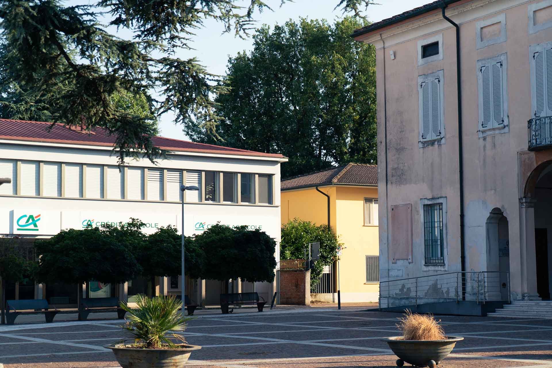 Scene di una piazza con alberi, edifici e panchine, in una giornata soleggiata.
