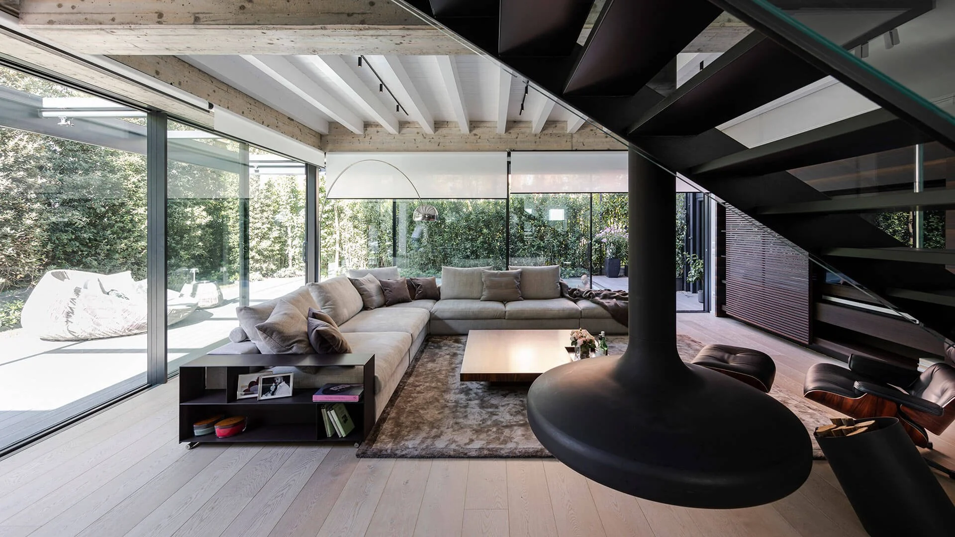 Salotto moderno con grande divano beige, tavolino, camino nero sospeso e scala a chiocciola in un ambiente luminoso con grandi finestre e pareti di vetro.